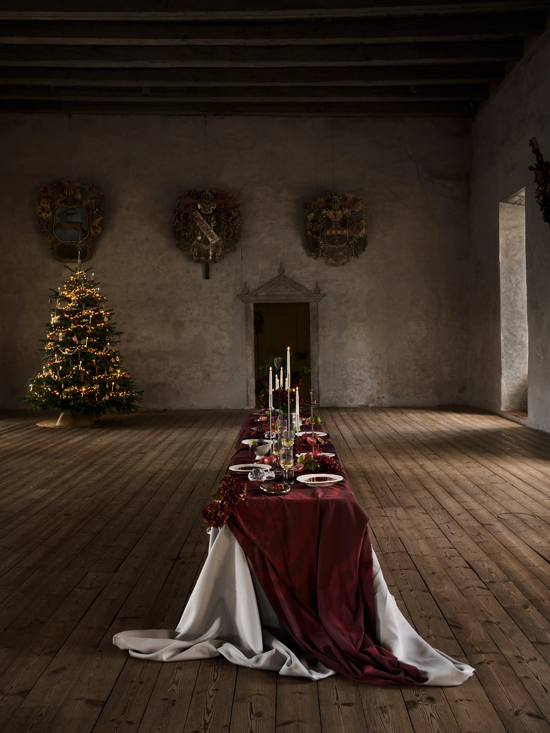 Lungo tavolo da cena di Natale con tovaglia rossa, candele, frutta e posti tavola, accanto a un albero di Natale illuminato in una vecchia sala.