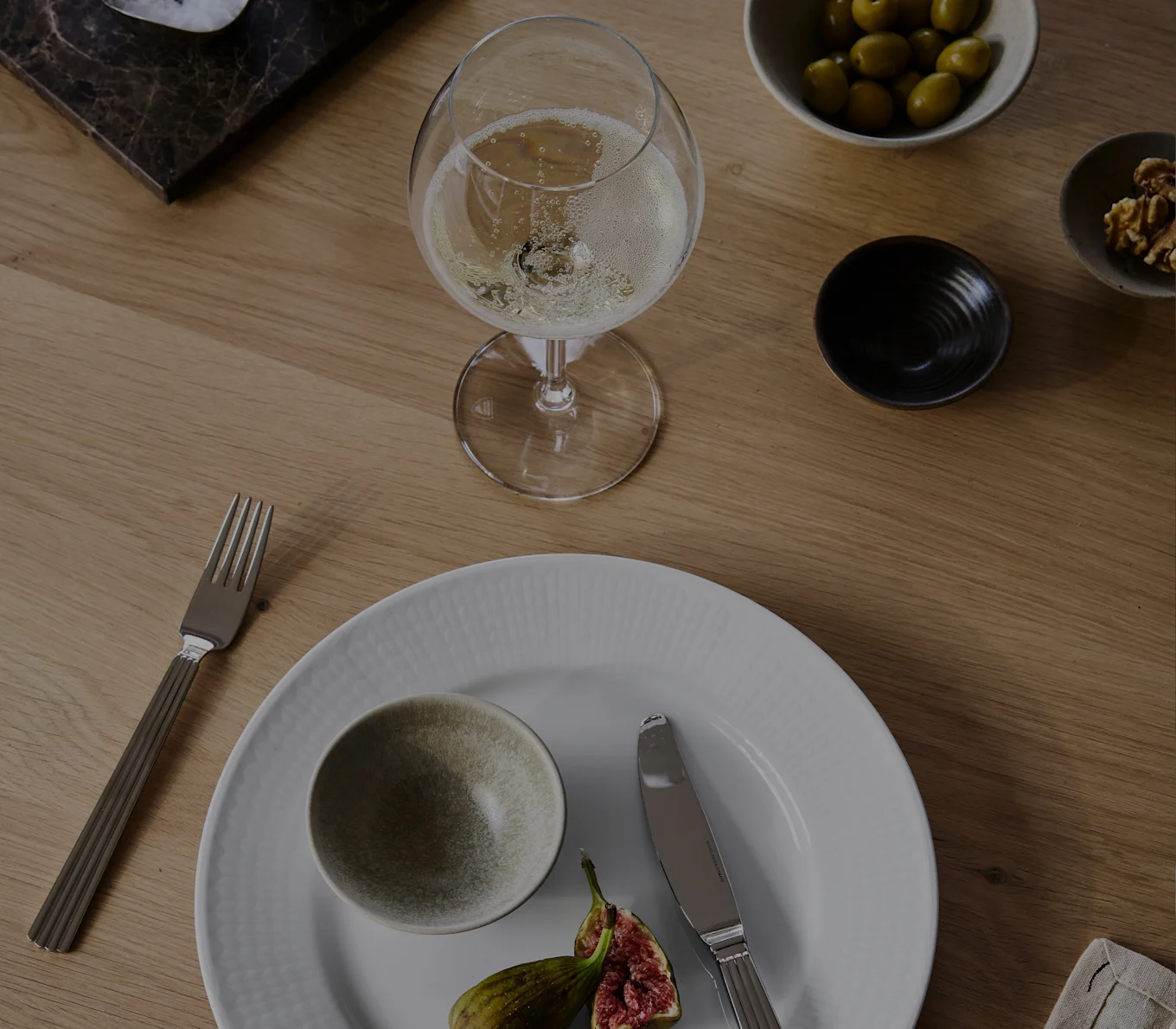 Un bicchiere di champagne, piatto bianco con ciotola e fichi, forchetta, coltello, olive e noci su un tavolo di legno.
