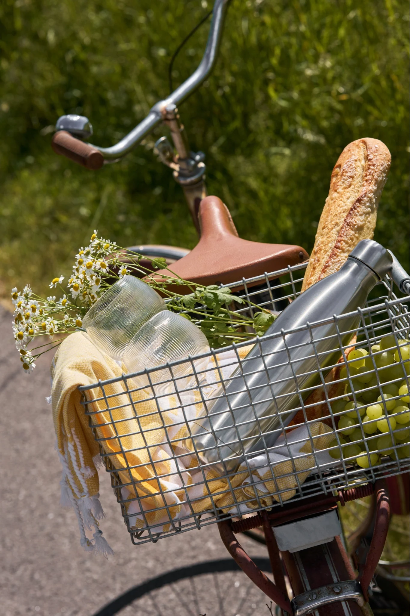 Bicicletta con cestino anteriore pieno di articoli da picnic: pane, uva verde, una borraccia d'argento, bicchieri, un asciugamano giallo e fiori.