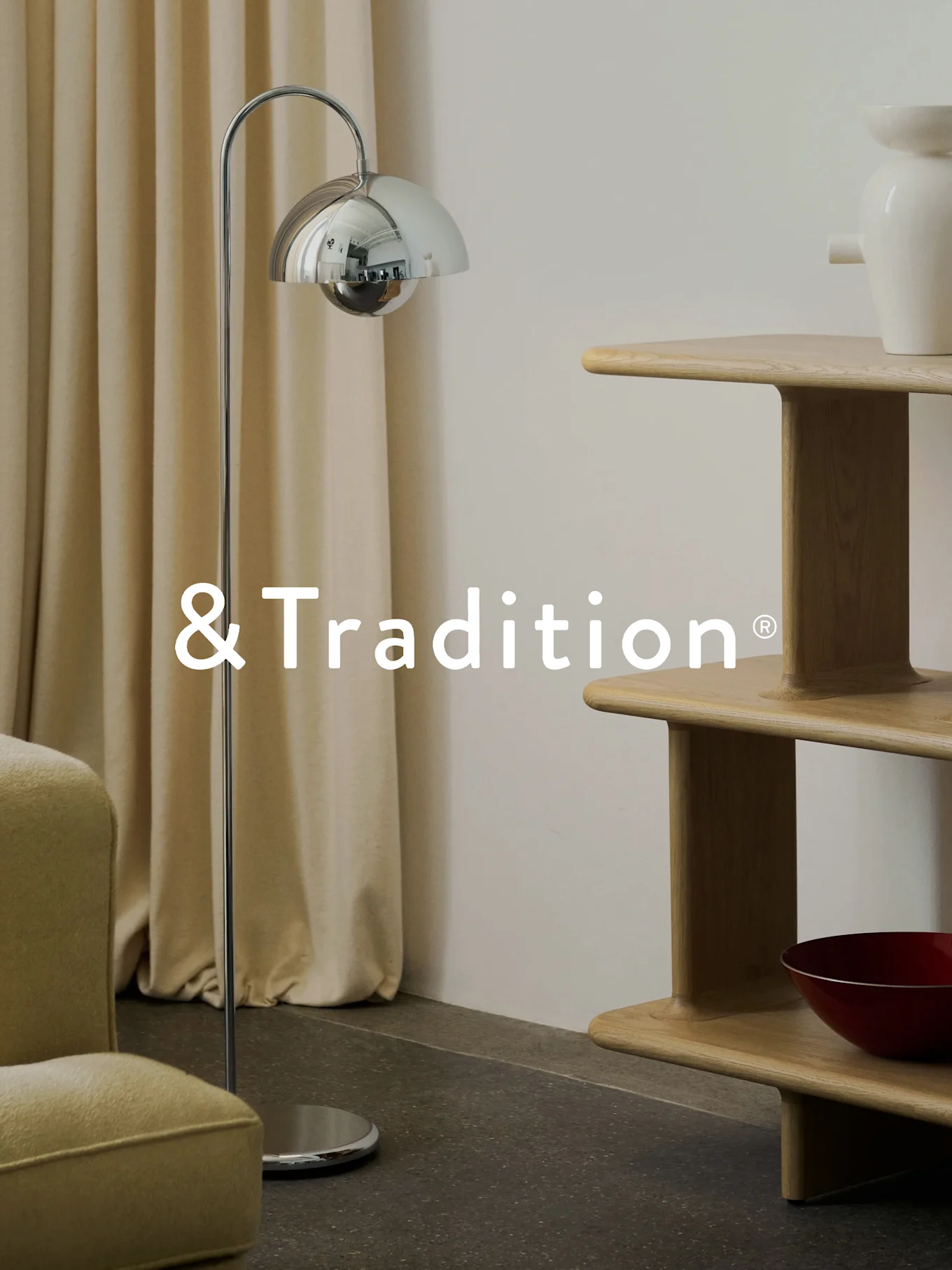 Un interno moderno con una lampada da terra cromata, una libreria a più livelli in legno chiaro e una poltrona. Logo &Tradition®.