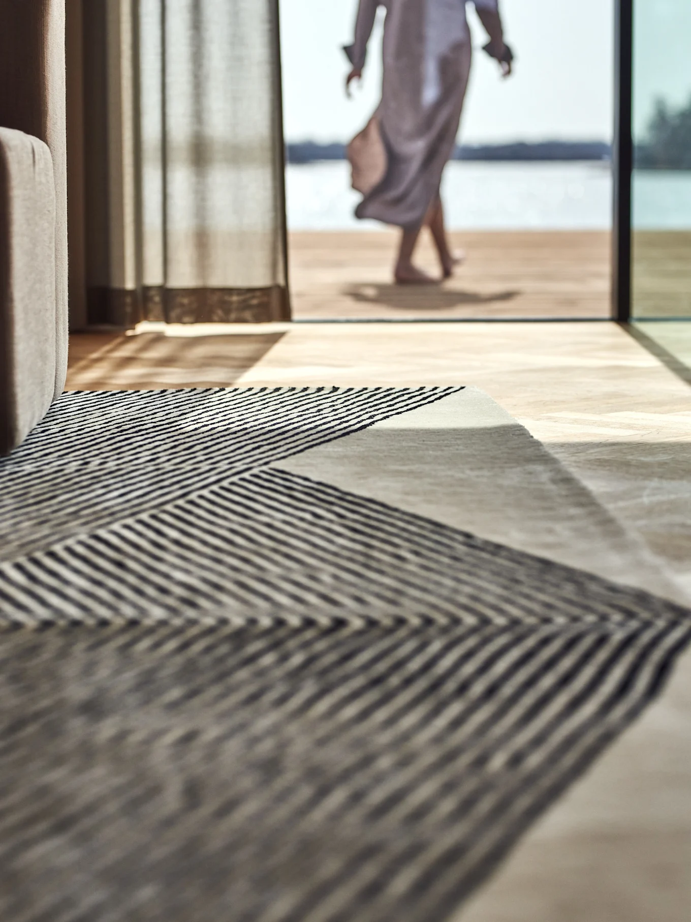 Tappeto a righe geometriche su pavimento in legno, con una persona che cammina verso un lago all'esterno.