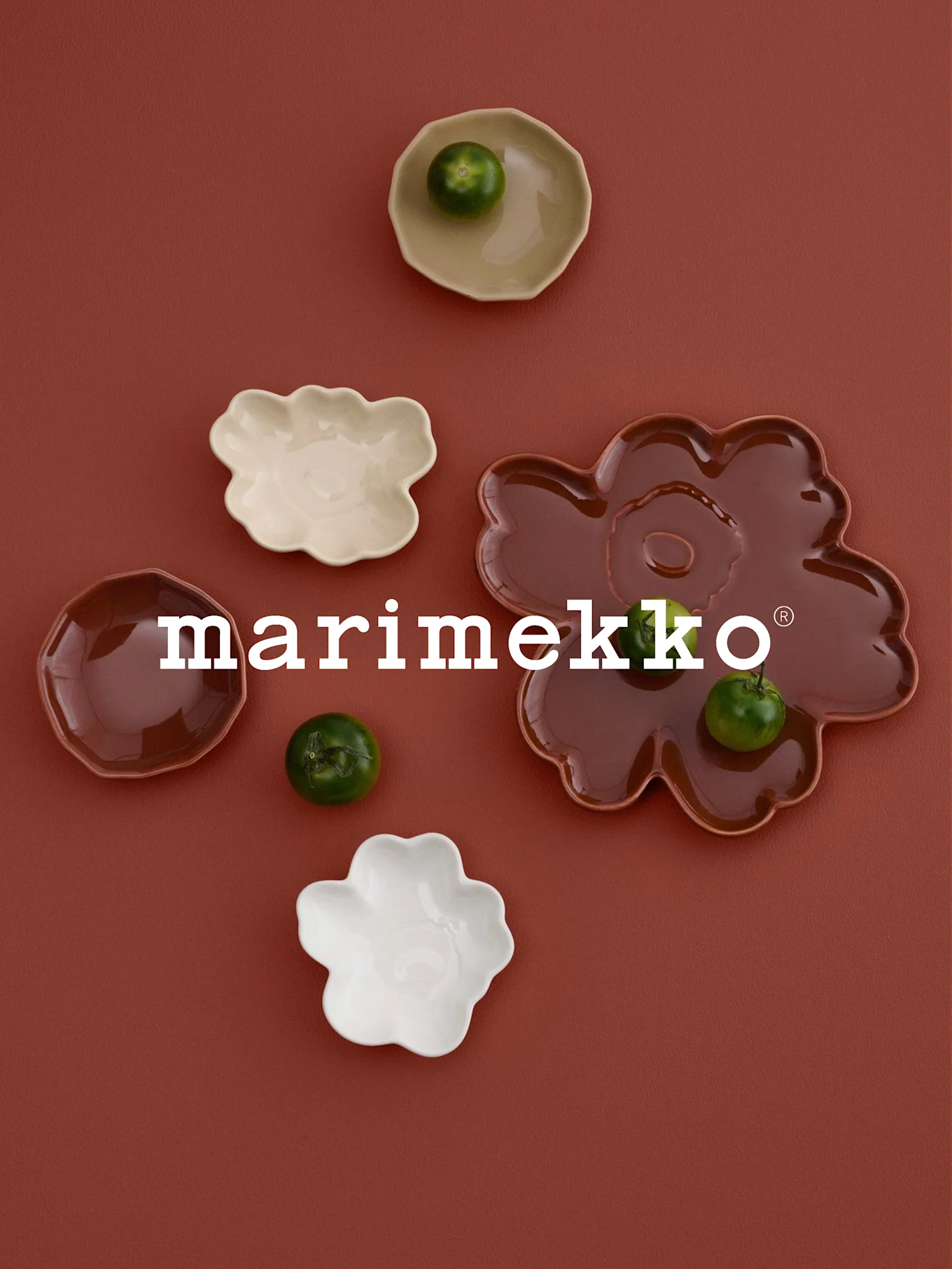 Logo bianco 'marimekko', piatti a forma di fiore (bianco, crema, marrone rossastro) con frutti verdi, su superficie color ruggine.