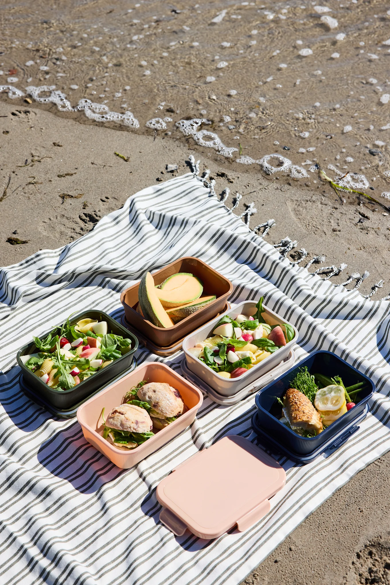 Cinque colorati porta pranzo aperti con insalate, panini e melone, più una scatola rosa chiusa, su una coperta a righe in spiaggia.