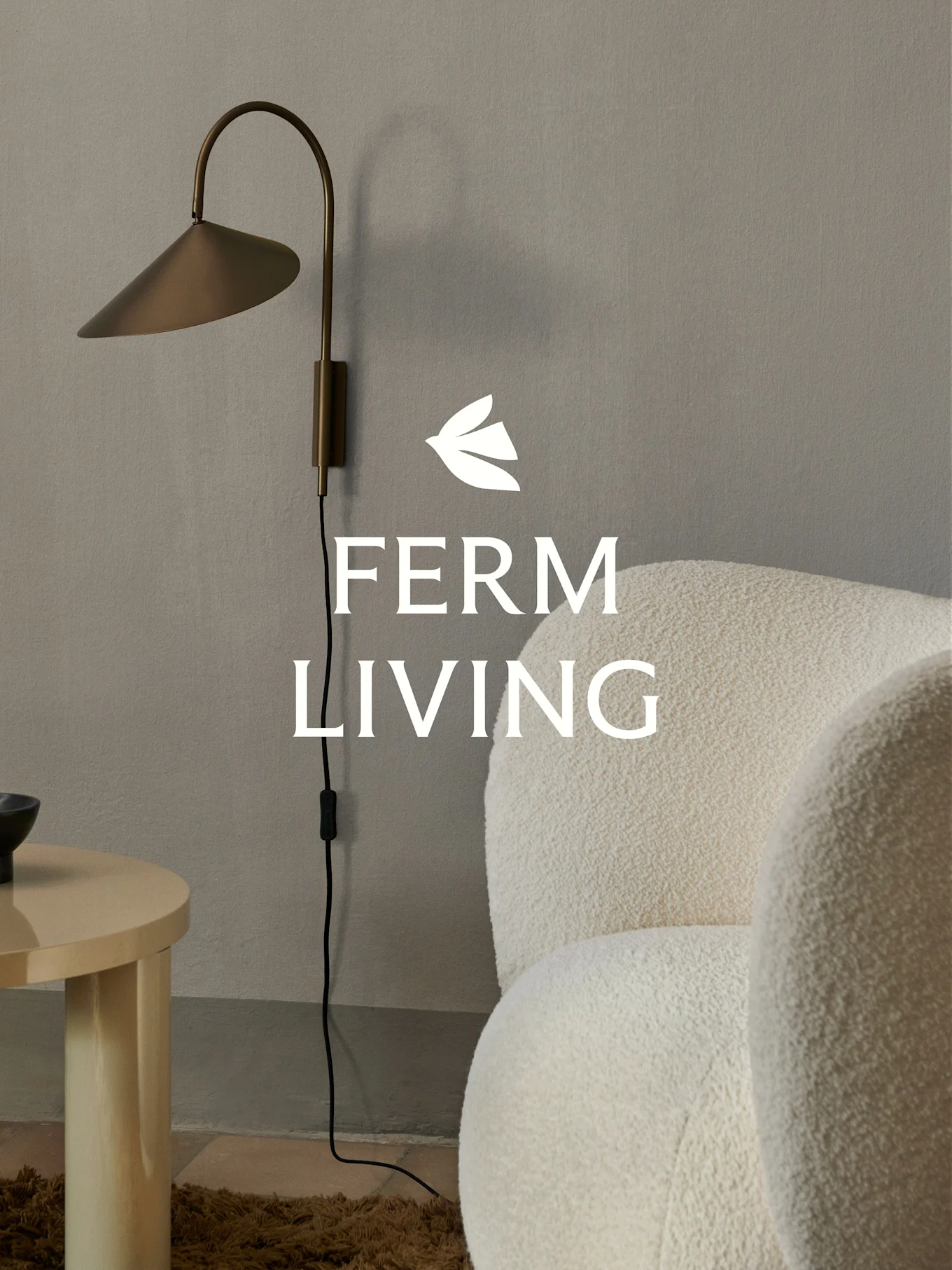 Logo Ferm Living su una lampada da parete in ottone, una poltrona bouclé crema e un tavolino in legno chiaro in un ambiente minimalista.