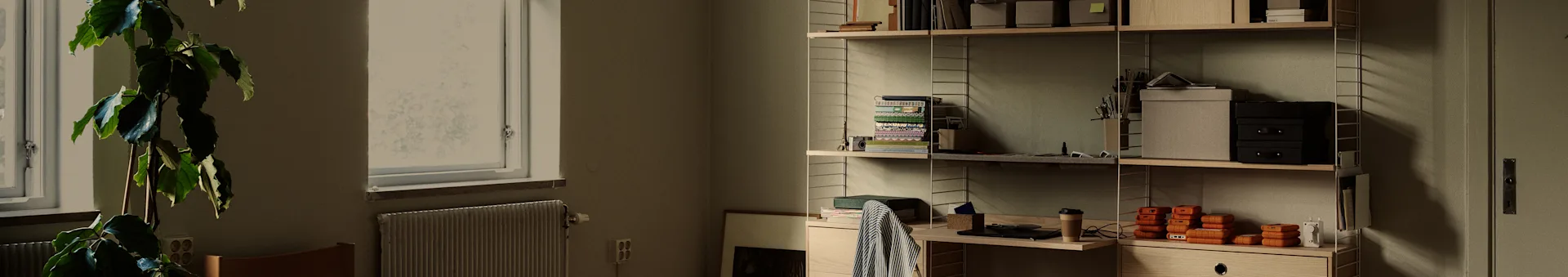 Un ordinato ufficio domestico con una grande pianta vicino a una finestra e una libreria in legno piena di libri, scatole e dischi rigidi portatili arancioni.