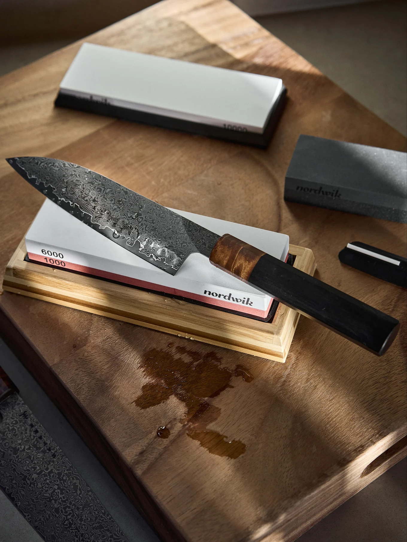Coltello da chef in Damasco su una pietra per affilare (grana 1000/6000) su un tagliere di legno con gocce d'acqua.