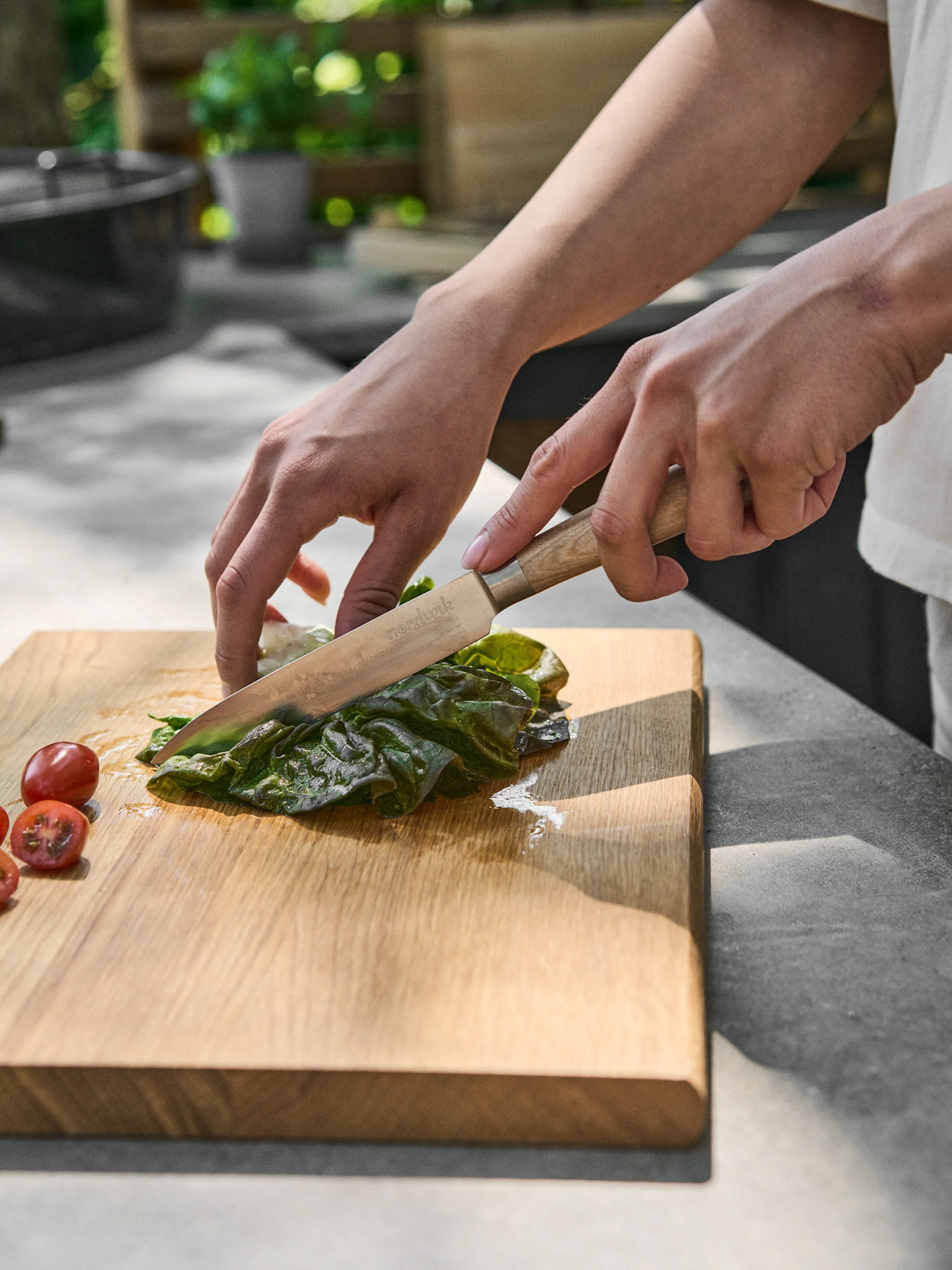 Mani che tagliano una foglia verde con un coltello su un tagliere di legno, accanto a pomodorini.