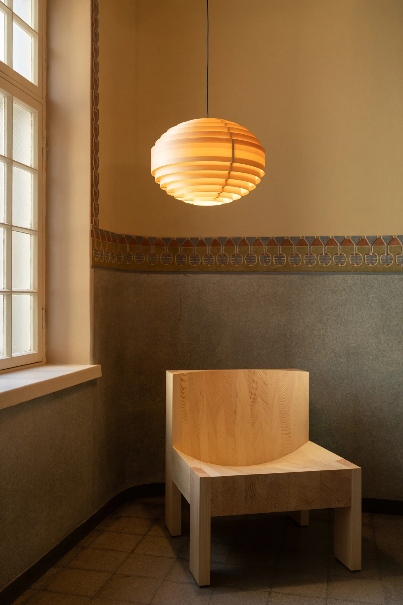 Lampada a sospensione a doghe di legno sopra una sedia in legno chiaro in una stanza con pareti decorate e finestra.