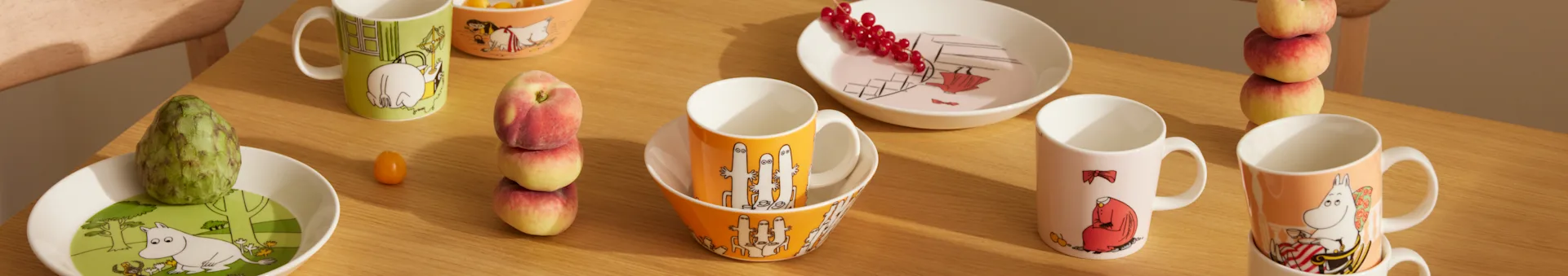Stoviglie Moomin in vari colori e design, con tazze, ciotole e piatti, insieme a frutta fresca su un tavolo di legno.