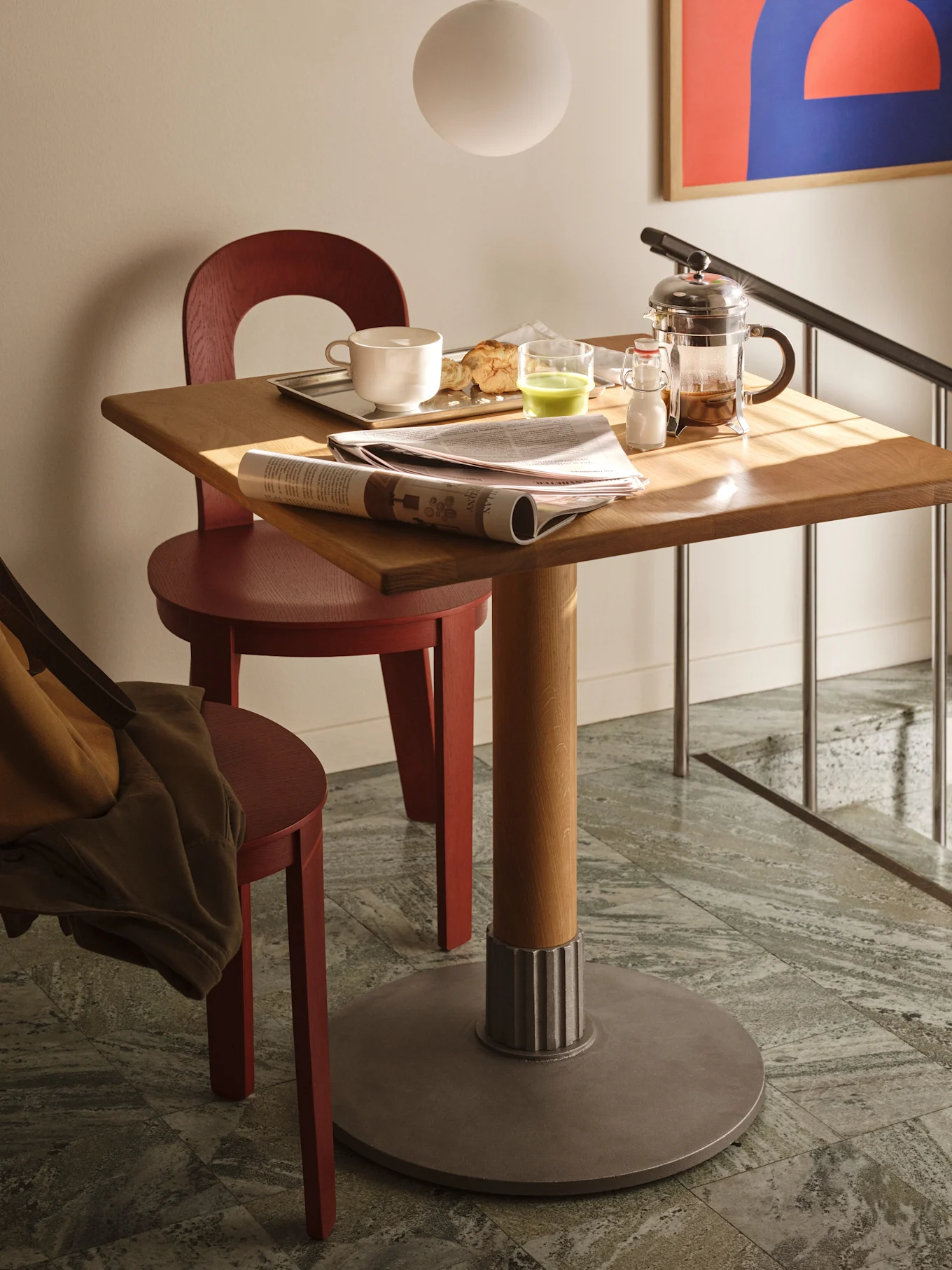 Un tavolo quadrato in legno con colazione, giornale e caffettiera francese, affiancato da due sedie rosse su piastrelle.
