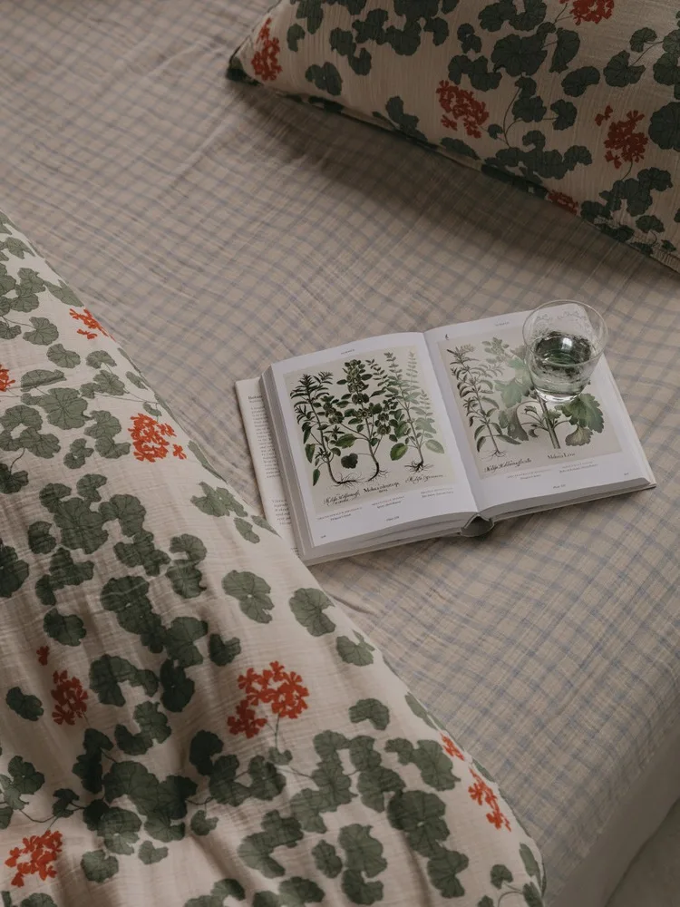 Un libro di botanica aperto e un bicchiere d'acqua su un letto con piumone a quadri e cuscini a fantasia floreale.