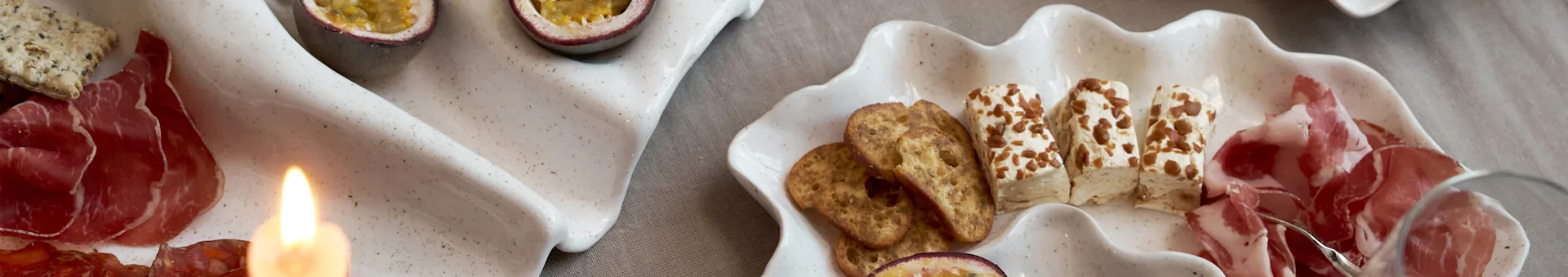 Un'esposizione di antipasti su due vassoi a forma di conchiglia con salumi, cracker, formaggio e frutto della passione, con una candela accesa.