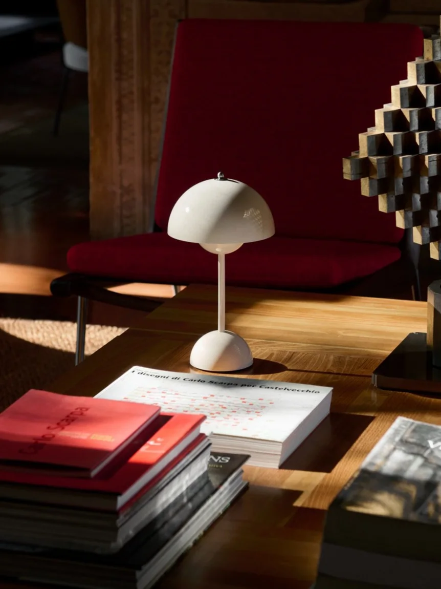 Una lampada da tavolo bianca a fungo, libri e una scultura geometrica su un tavolino da caffè in legno, con una poltrona rossa dietro.