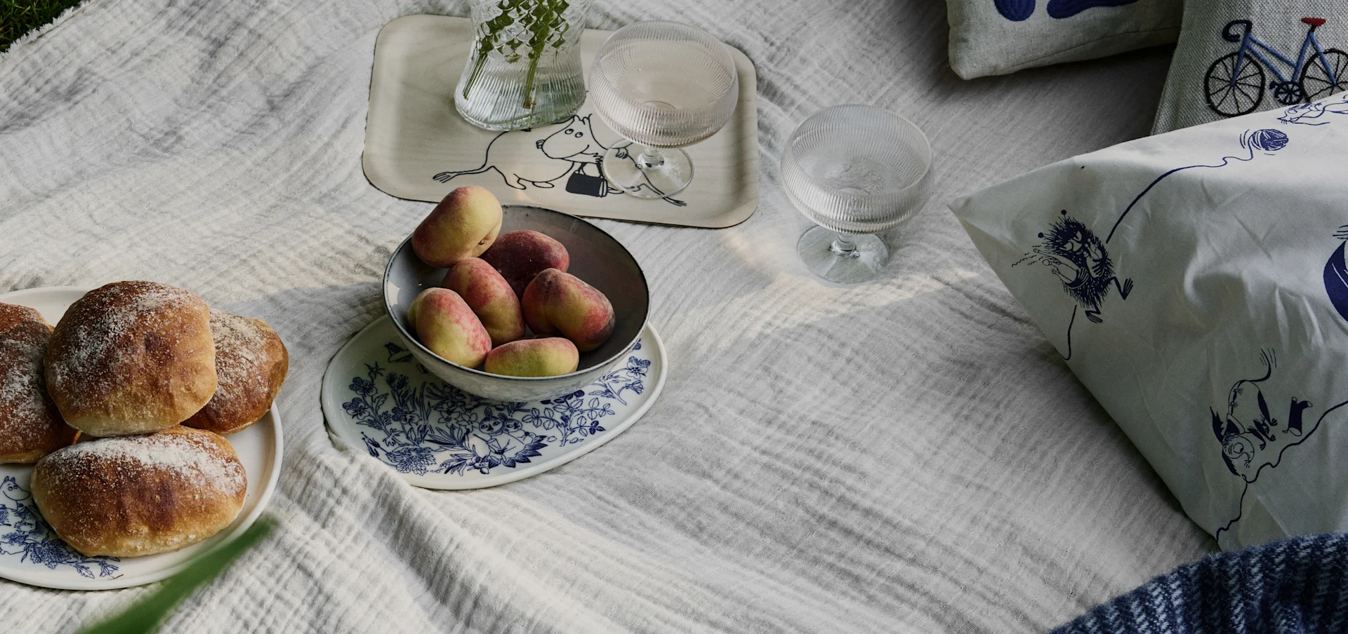 Scena di picnic su una coperta bianca con panini, pesche, bevande e cuscini con motivi Moomin e bicicletta.