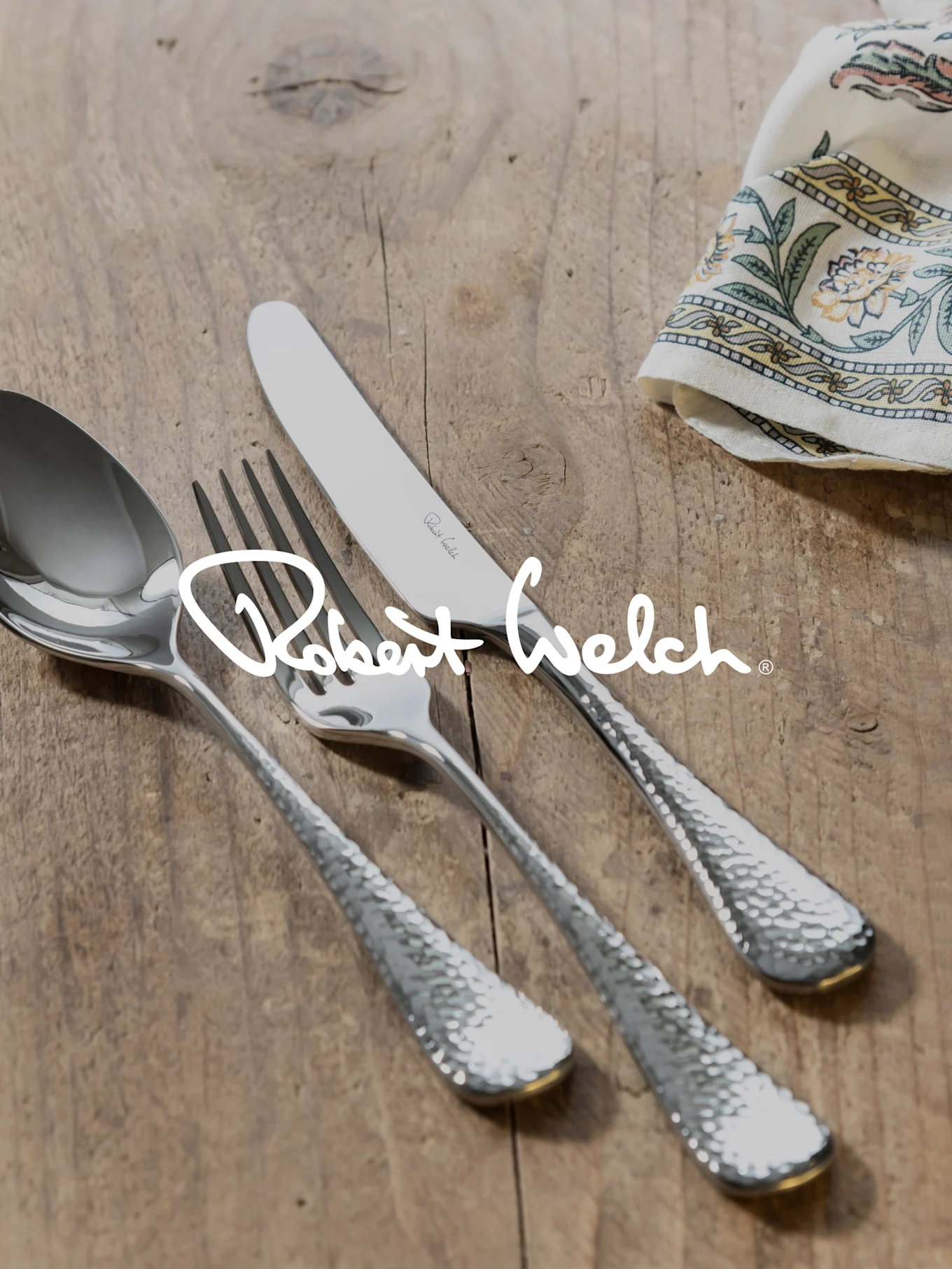 Logo Robert Welch su posate argentate martellate (cucchiaio, forchetta, coltello) su legno con panno fantasia.