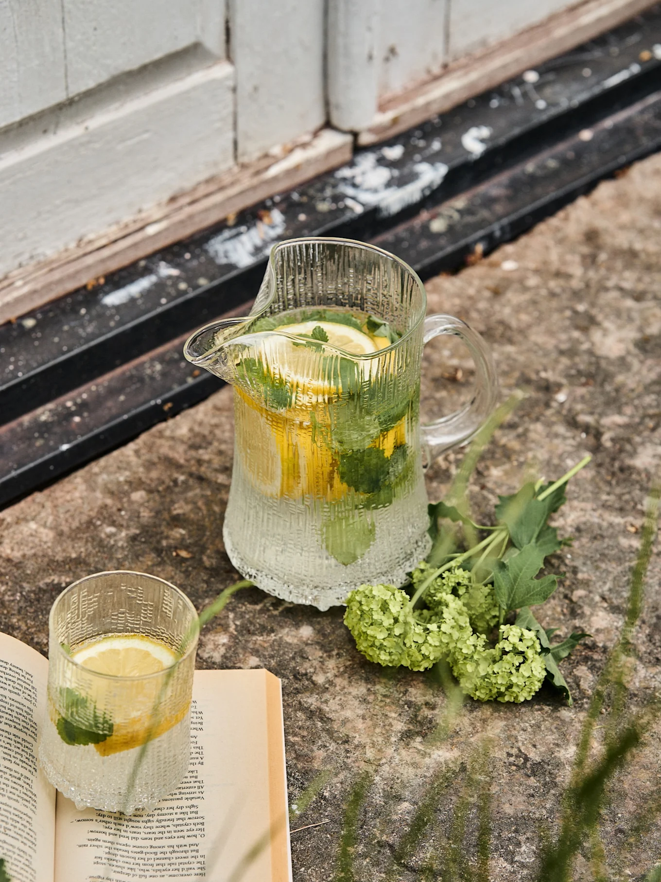 Brocca e bicchiere in vetro rigato con acqua, limone e menta, accanto a un libro aperto e fiori verdi su pietra.