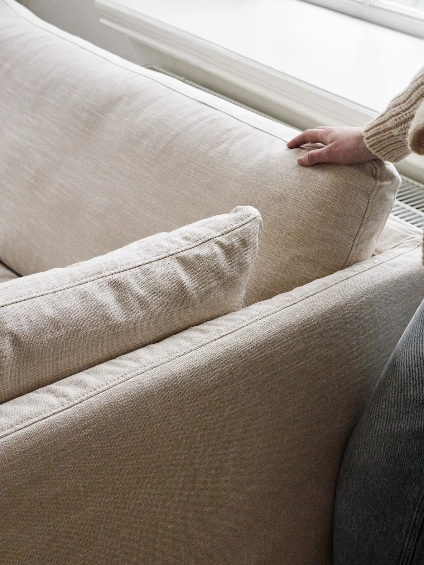 Una mano con un maglione a maglia grossa preme delicatamente il cuscino posteriore di un divano beige chiaro.
