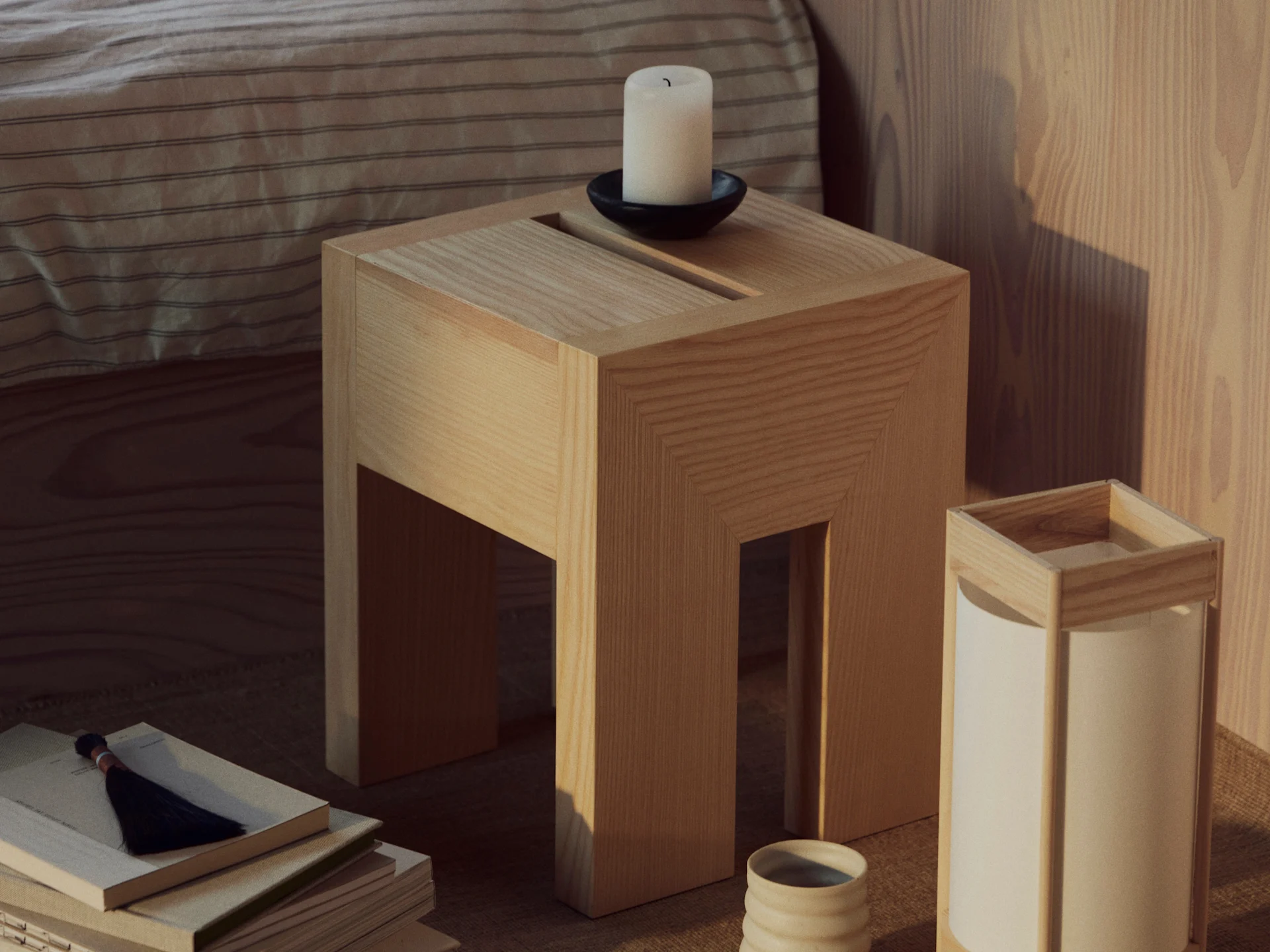 Un tavolino in legno con una candela bianca in un portacandele nero, una pila di libri con una nappa e una lampada con una tazza in ceramica.