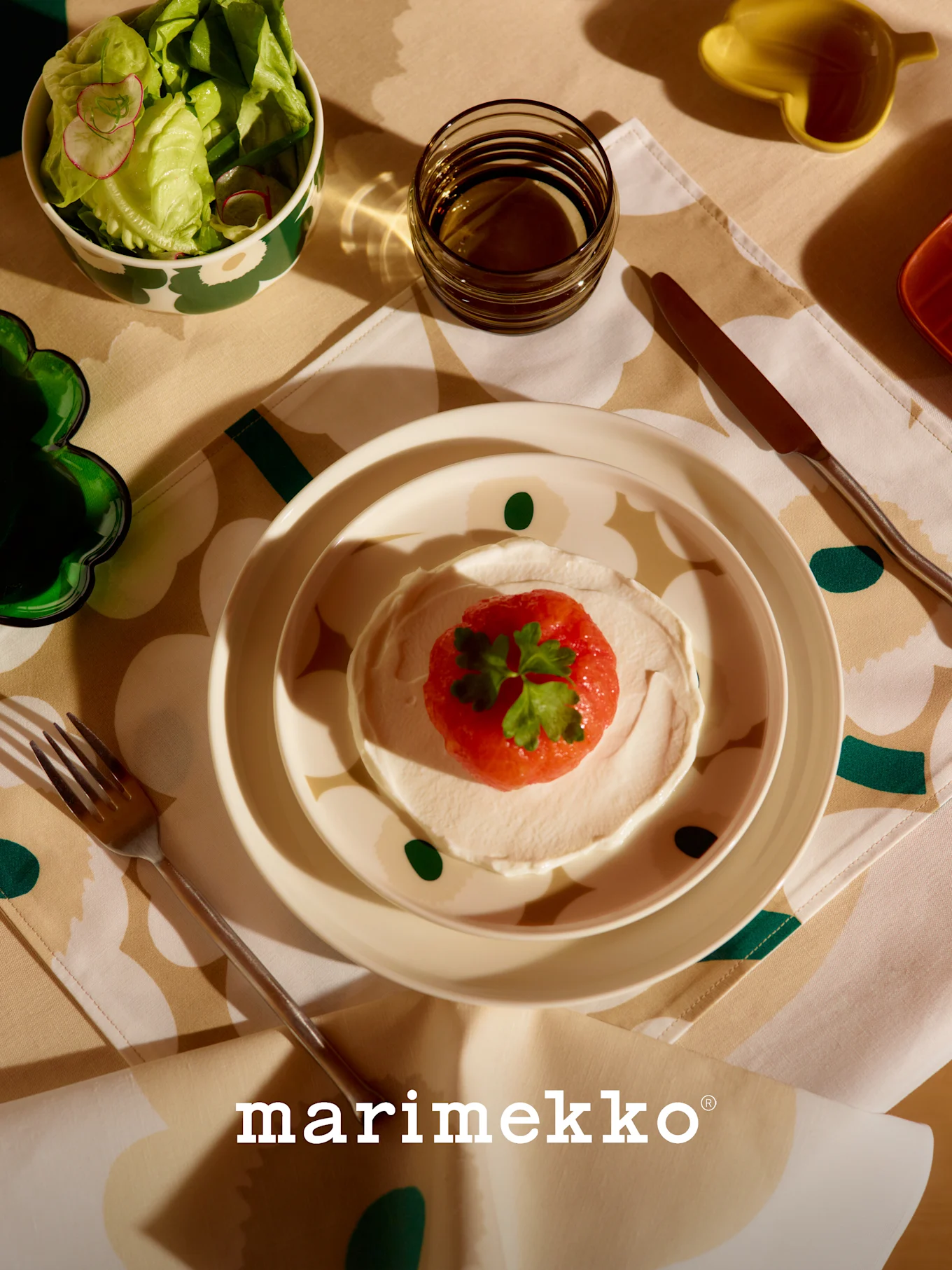 Tavola Marimekko con insalatiera a motivi, piatto di frutta rossa e crema, bicchiere e posate su tovaglia floreale.