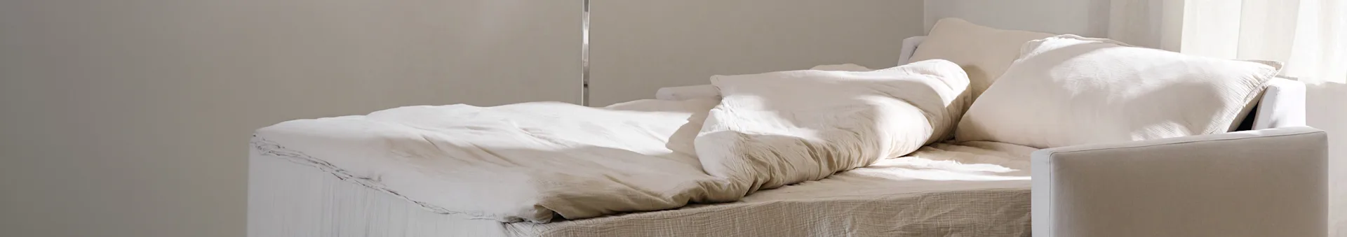 Un letto con lenzuola e cuscini di colore chiaro stropicciati, illuminato delicatamente dalla luce naturale.