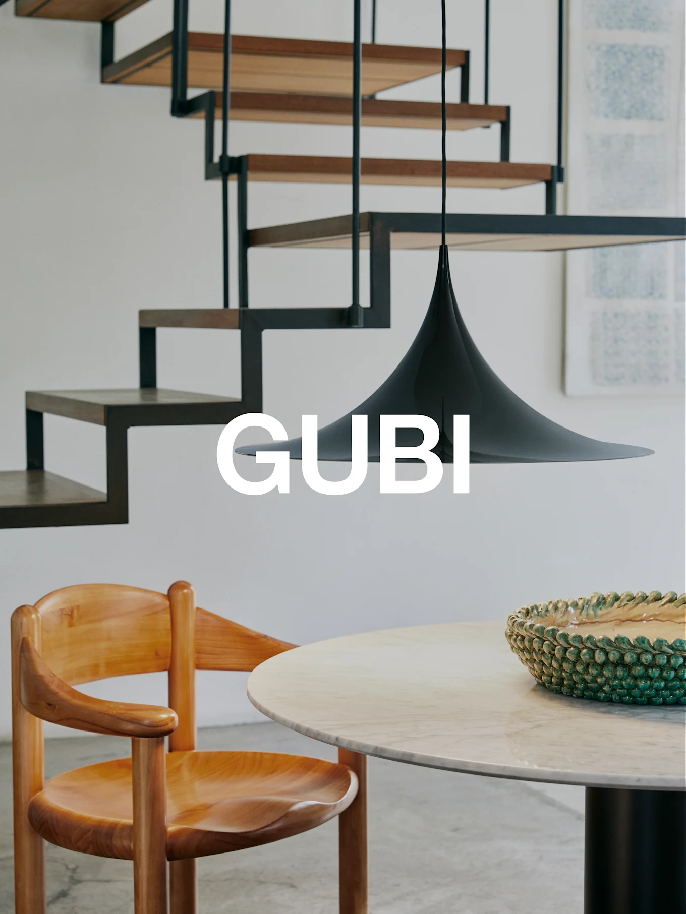 Logo GUBI in un interno moderno con sedia in legno, tavolo in marmo, ciotola verde, lampada a sospensione nera e scala moderna.