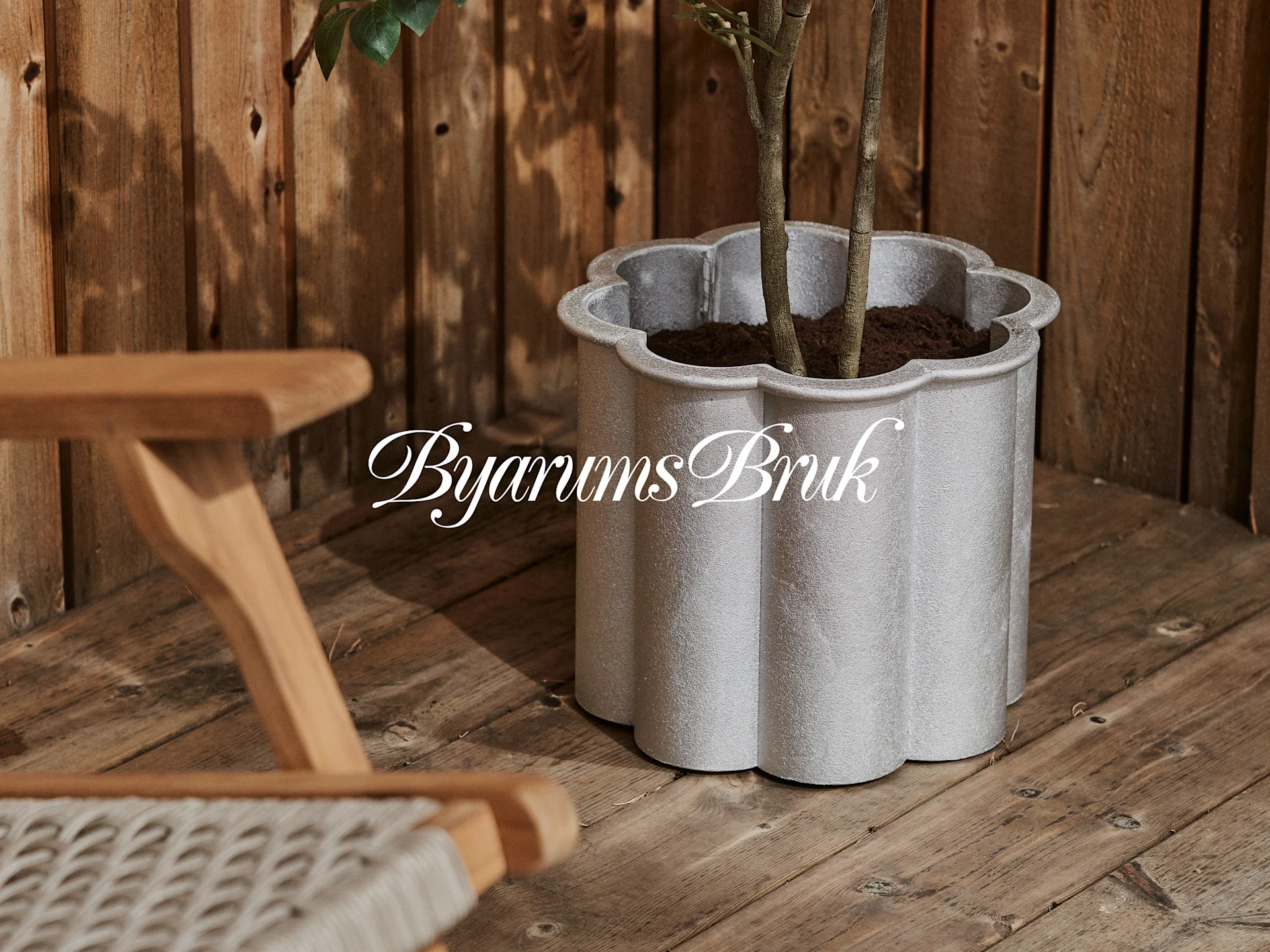 Una fioriera smerlata grigio-argentata con una pianta su un ponte di legno, accanto a una sedia di legno. Logo 'Byarums Bruk'.