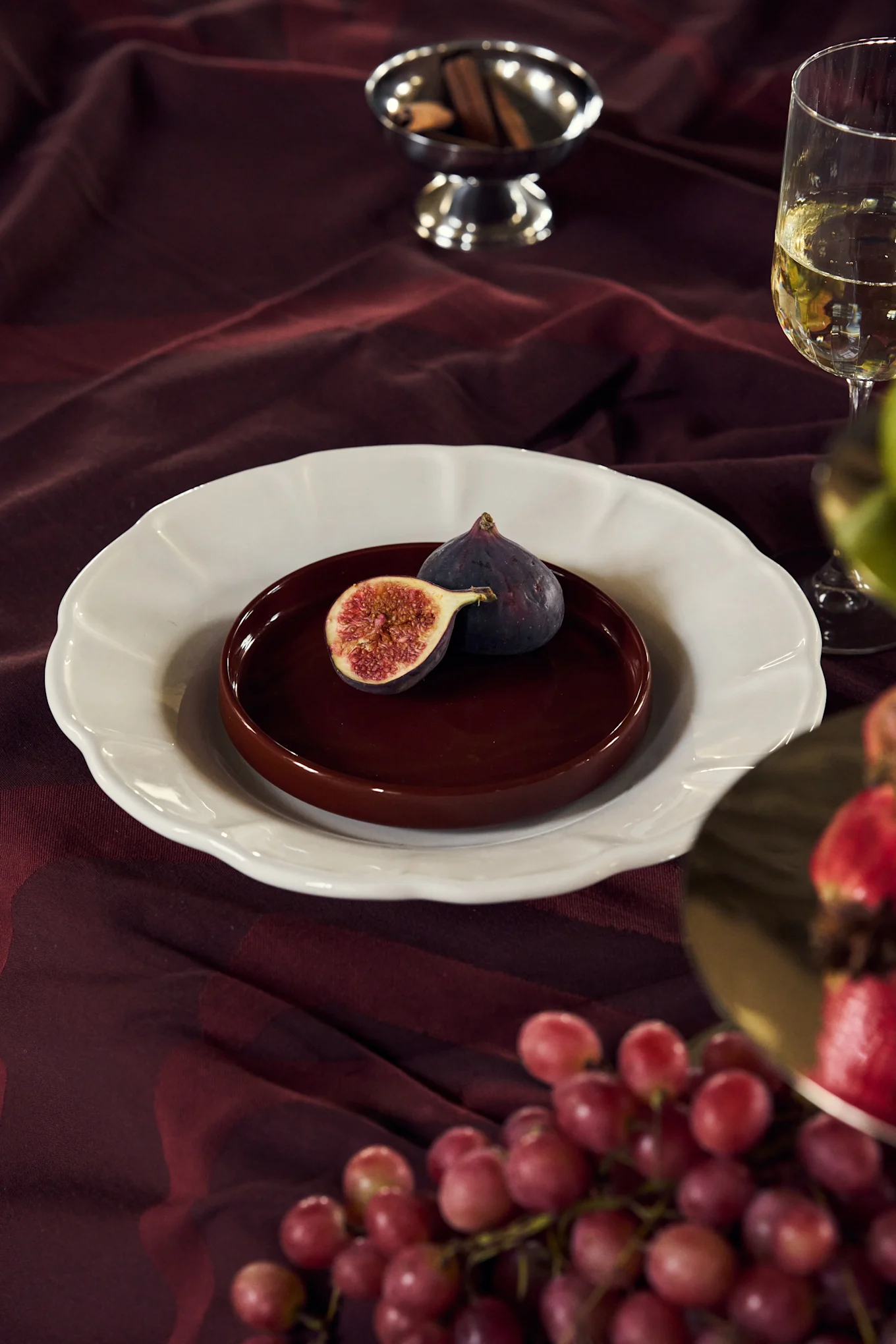 Fichi su un piatto sopra una tovaglia bordeaux con vino, uva e bastoncini di cannella.