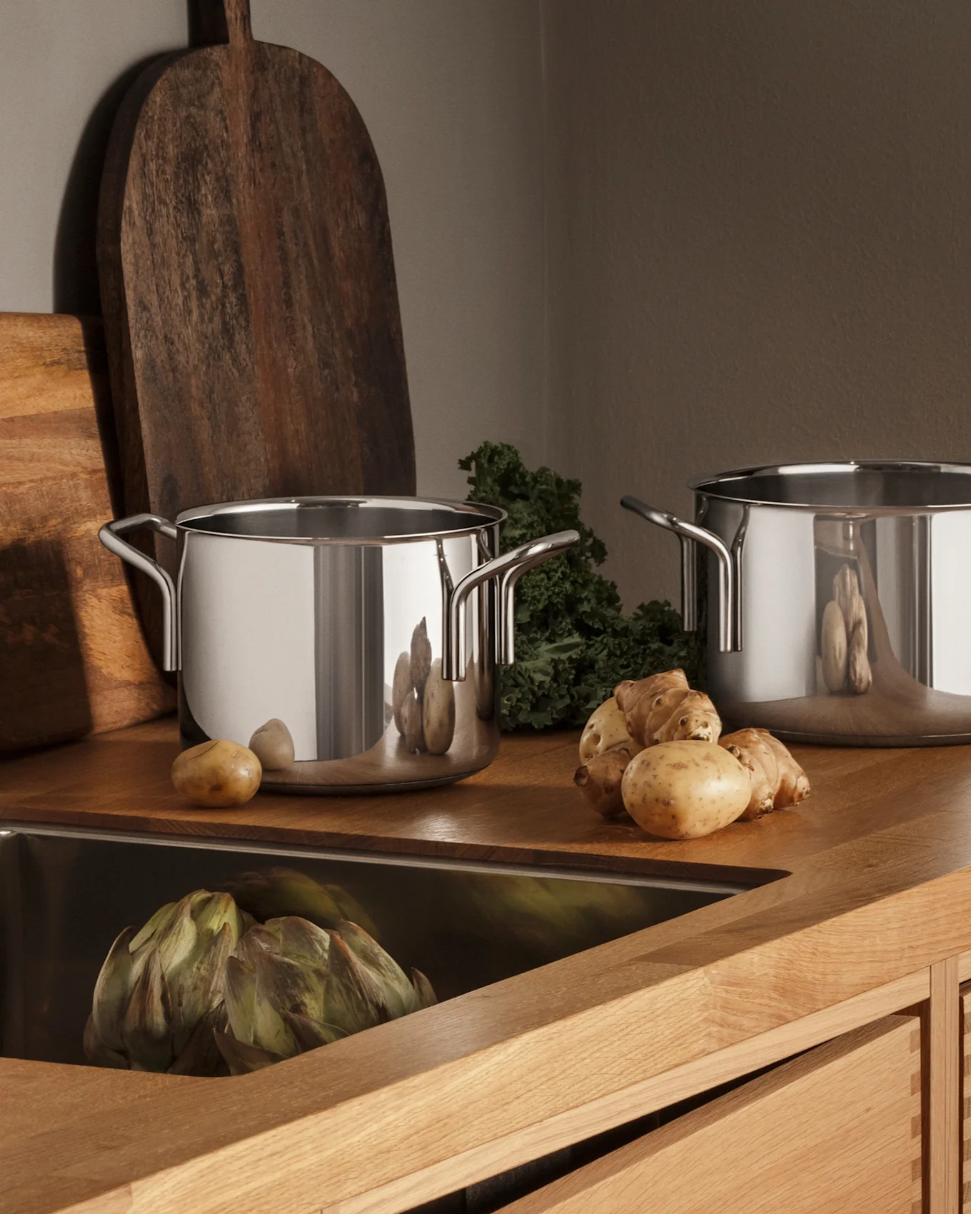 Cucina moderna con bancone in legno, lavello in acciaio inossidabile, rubinetto, tre pentole e prodotti freschi come patate e carciofi.