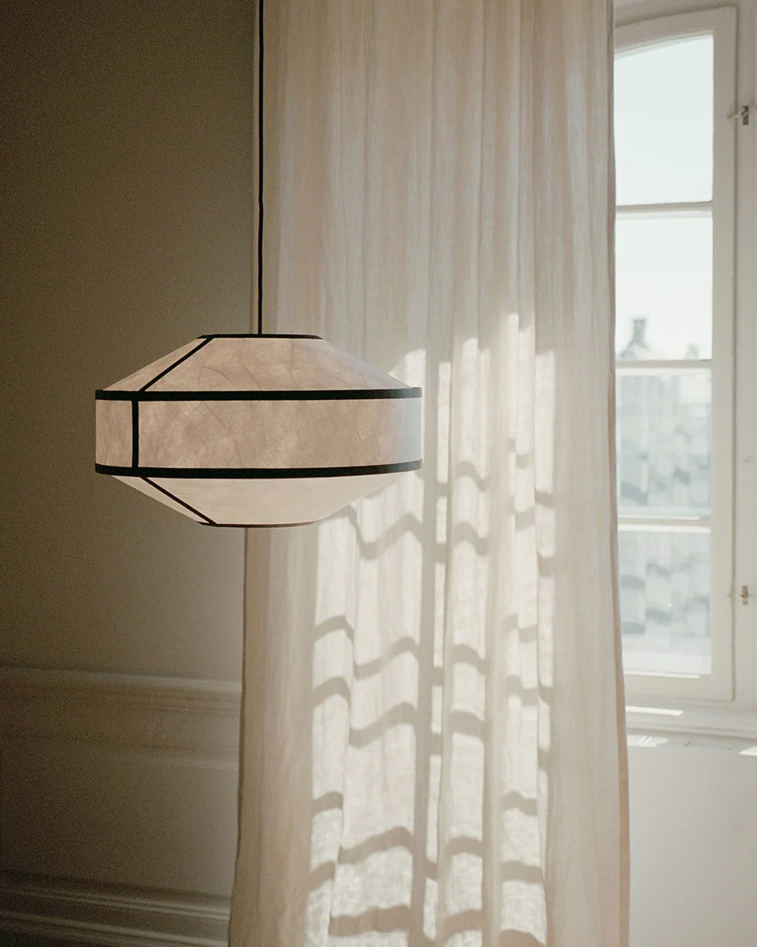 Una lampada a sospensione geometrica in carta con cornici scure pende accanto a una tenda trasparente e una finestra illuminate dal sole.