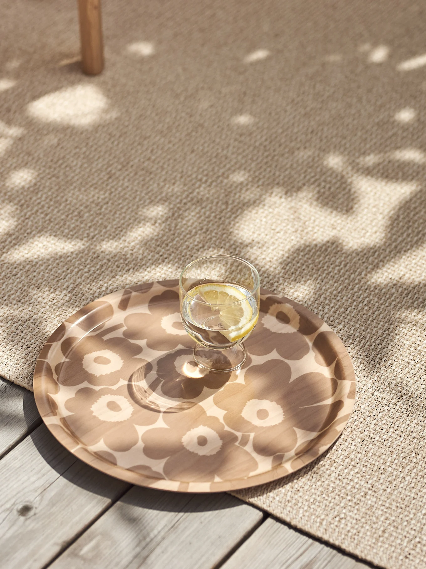 Bicchiere d'acqua e limone su un vassoio beige con motivo floreale su un tappeto strutturato con luce solare a chiazze.