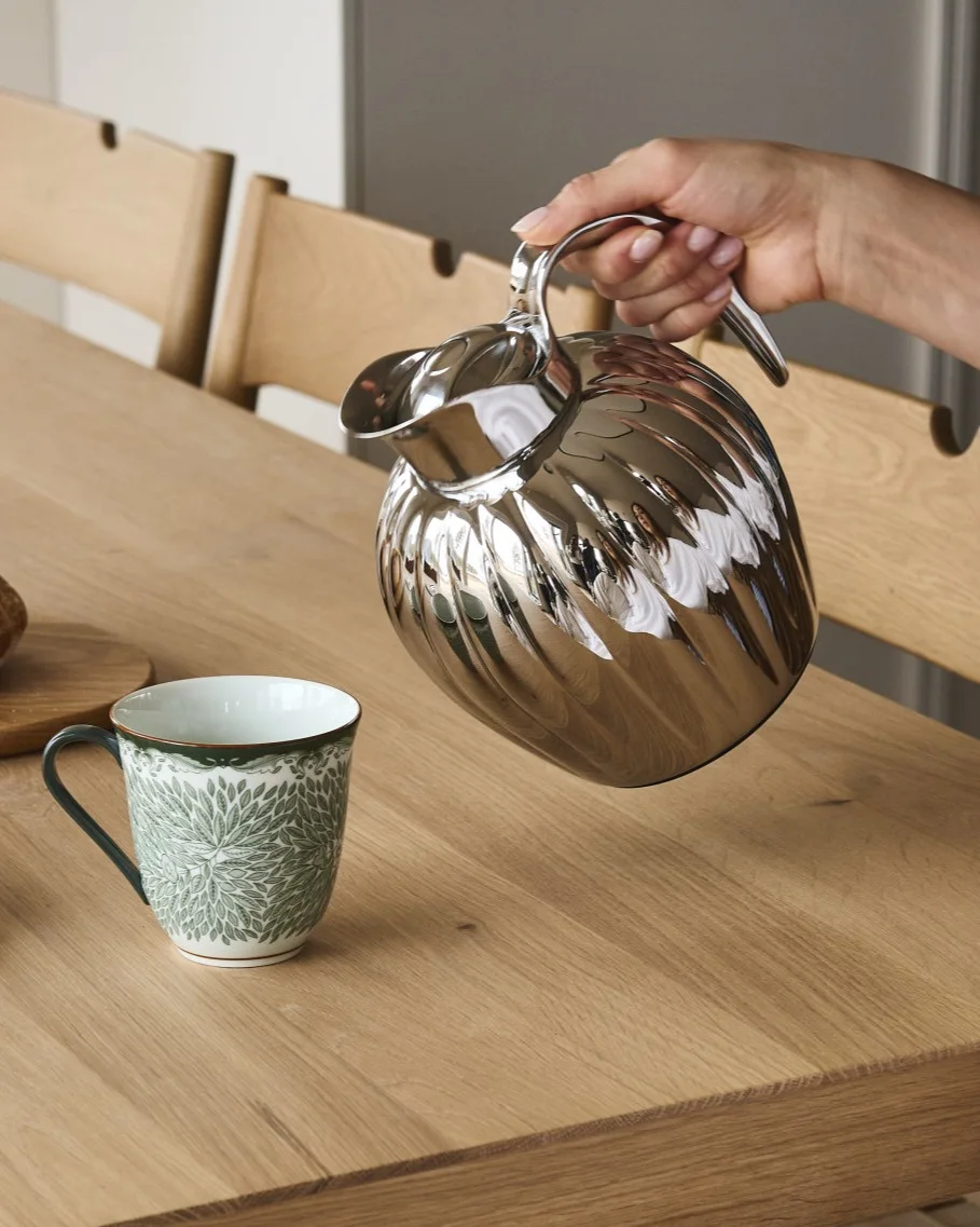 Una mano versa da una caraffa argentata a coste in una tazza bianca con motivo a foglie verdi su un tavolo di legno.