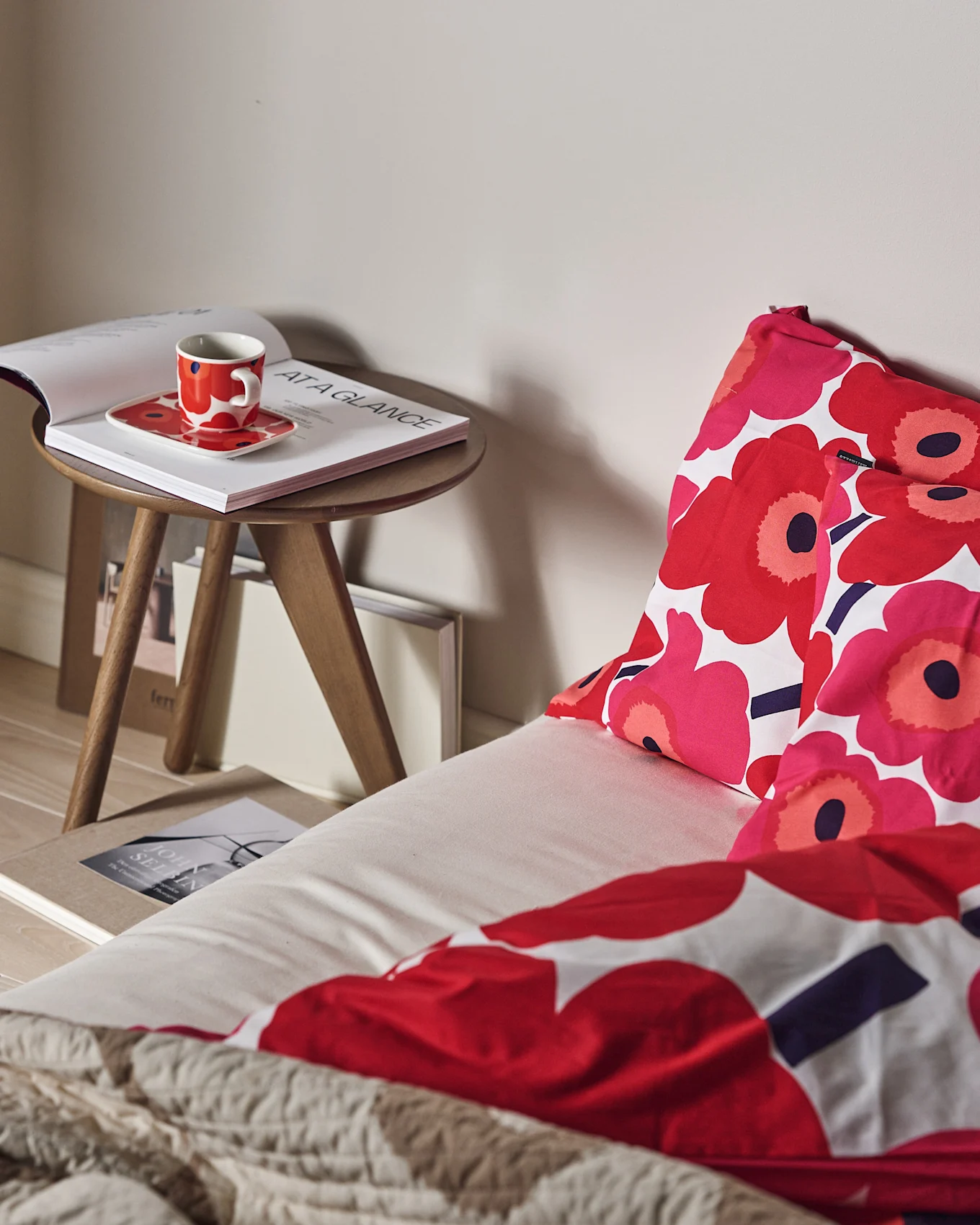 Un letto con biancheria da letto floreale rossa e rosa e un plaid beige testurizzato. Una tazza rossa e un libro aperto su un tavolino in legno.