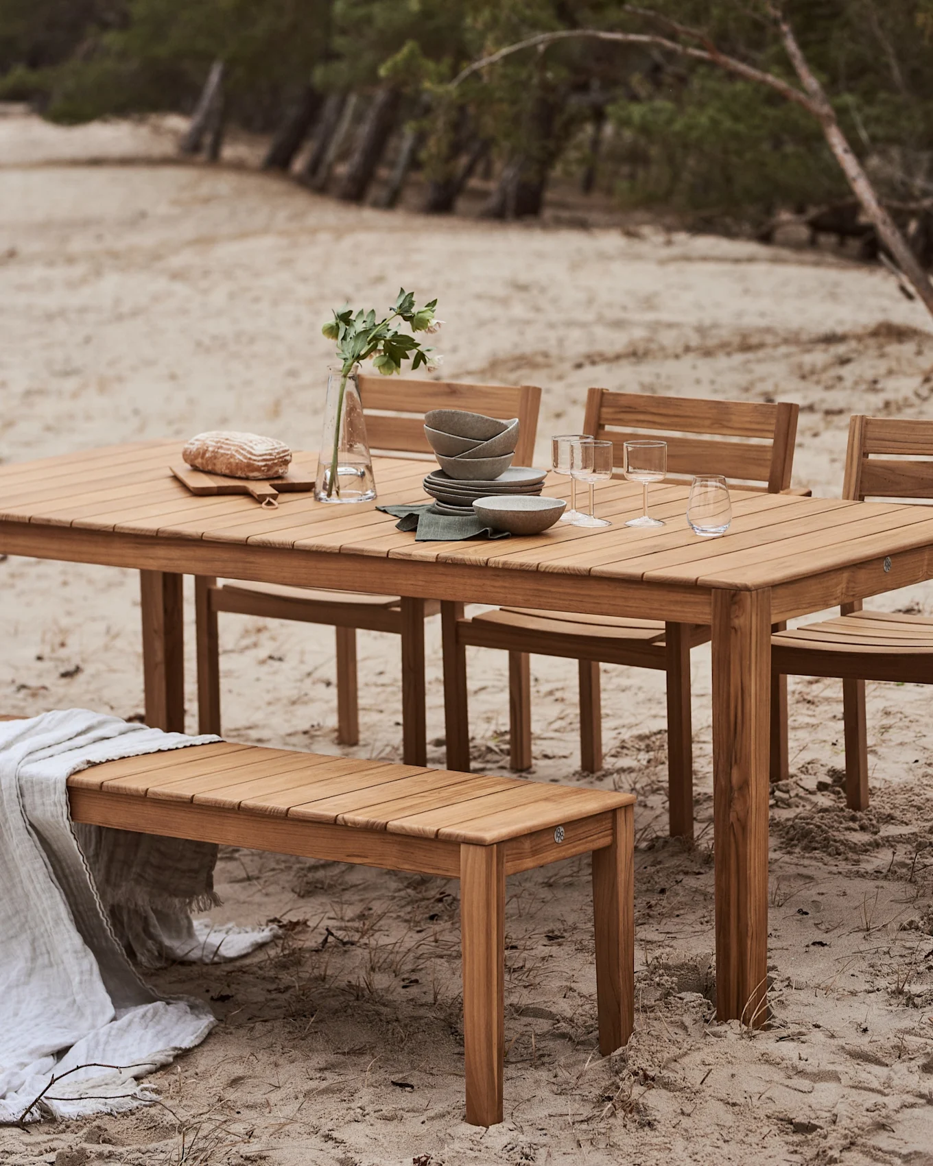 Set da pranzo in legno per esterni con tavolo e panca su una spiaggia sabbiosa. Il tavolo è apparecchiato con piatti, bicchieri, pane e un vaso di fiori.
