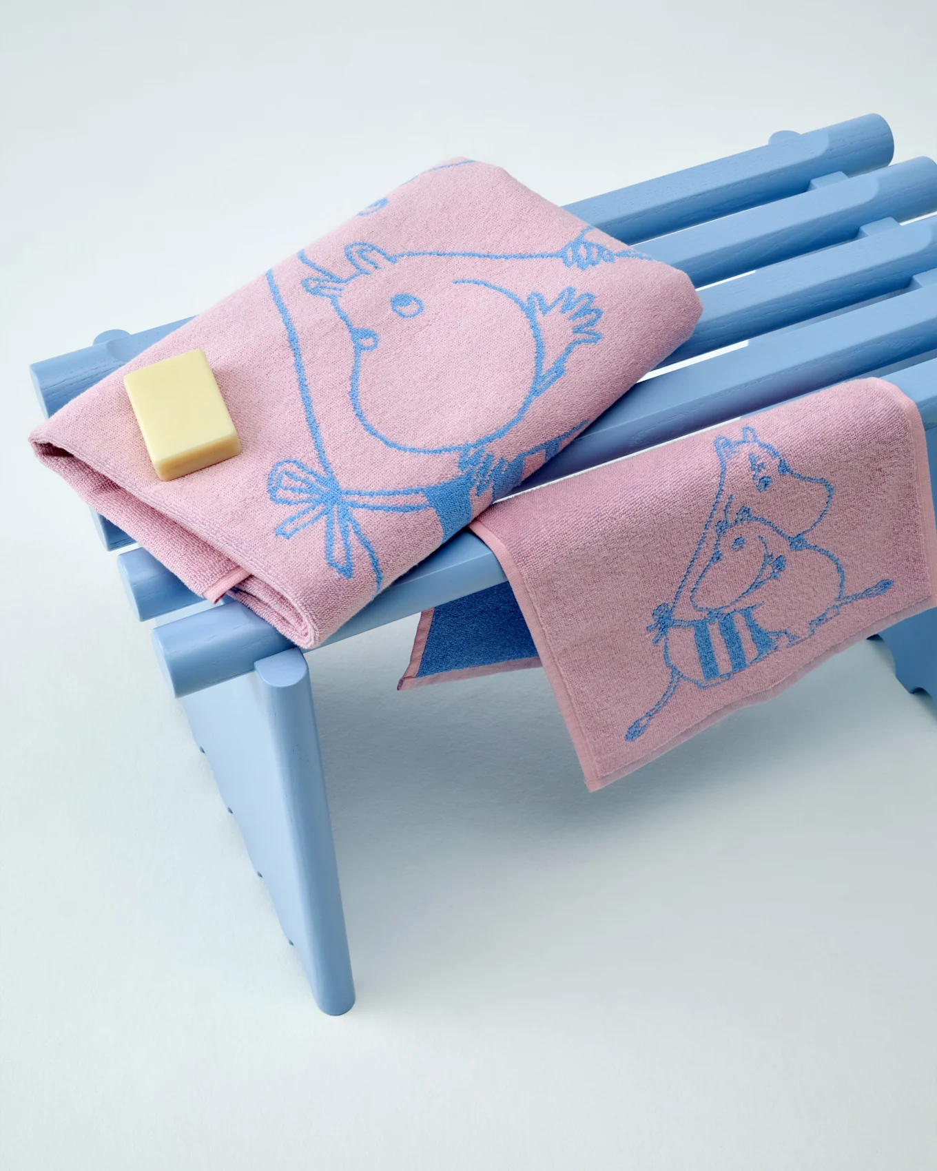 Due asciugamani Moomin rosa con fantasie blu, uno piegato con una saponetta, su una panca azzurra.
