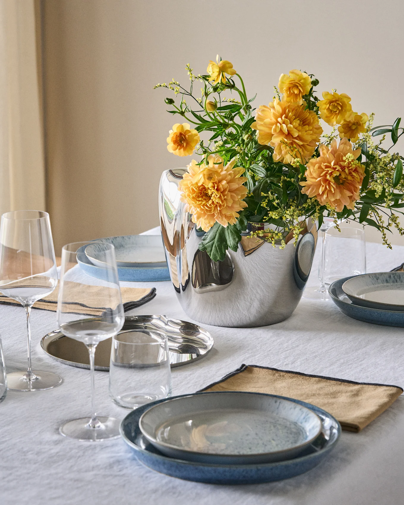 Un tavolo da pranzo apparecchiato con piatti blu, tovaglioli beige, bicchieri trasparenti e un vaso d'argento con fiori giallo-arancio su una tovaglia bianca.