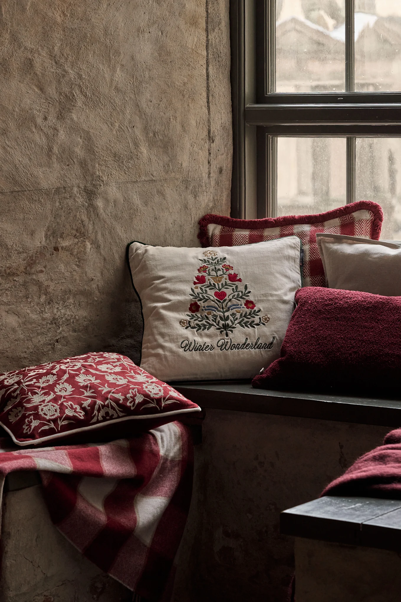 Accogliente nicchia finestra con cuscini festivi, incluso uno con albero di Natale ricamato e 'Winter Wonderland', e una coperta a quadri rossa.