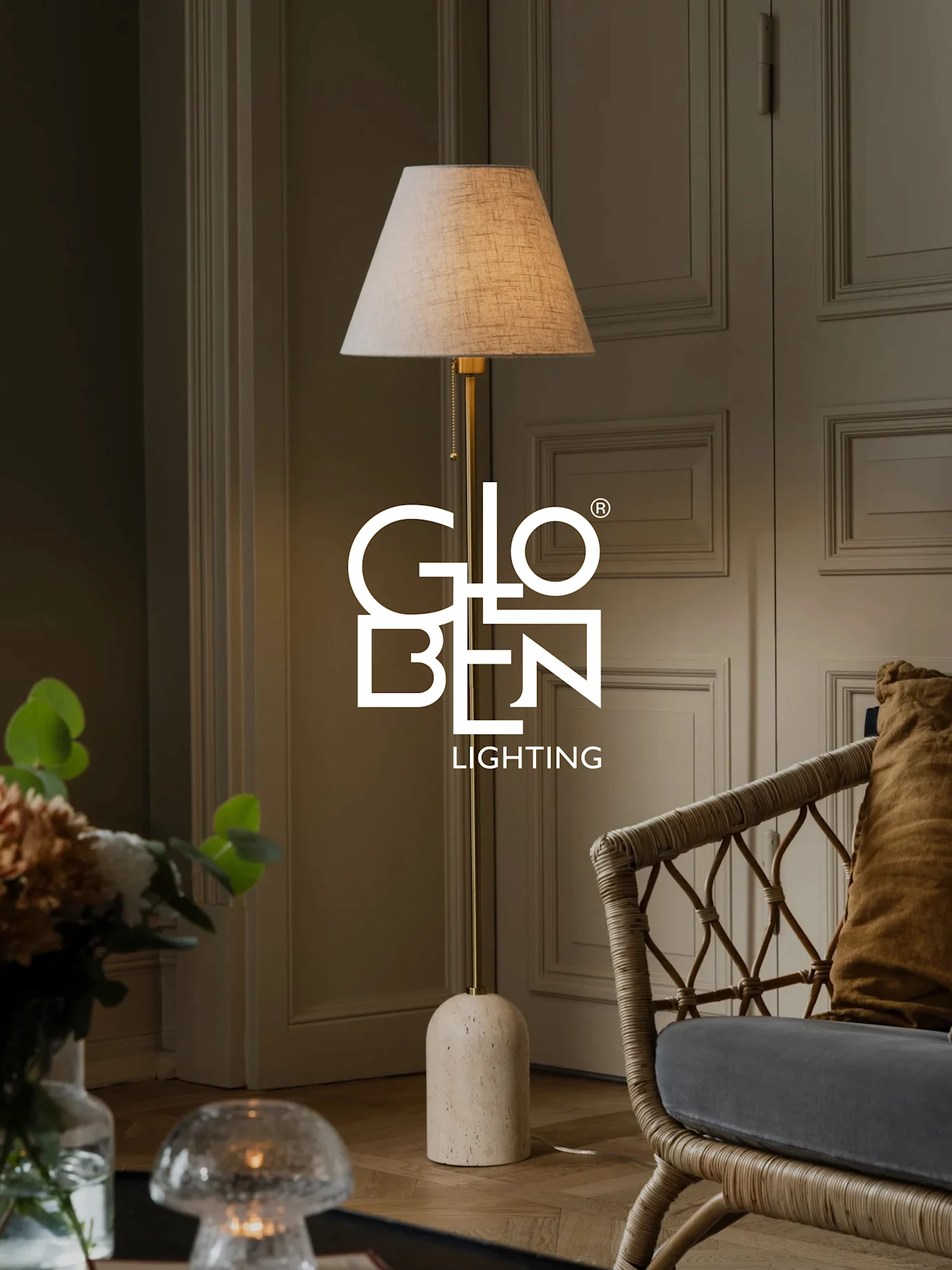 Una lampada da terra con paralume beige, stelo in ottone e base in pietra arrotondata accanto a una sedia in vimini.