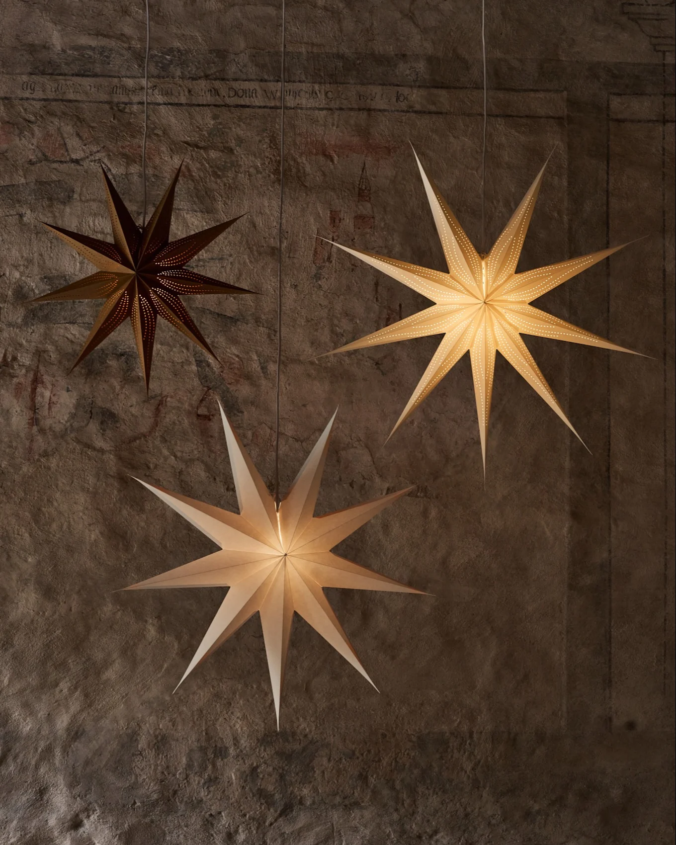 Tre lanterne di carta a forma di stella pendono su una parete ruvida. Una scura ha piccole perforazioni, due più luminose illuminano la scena.