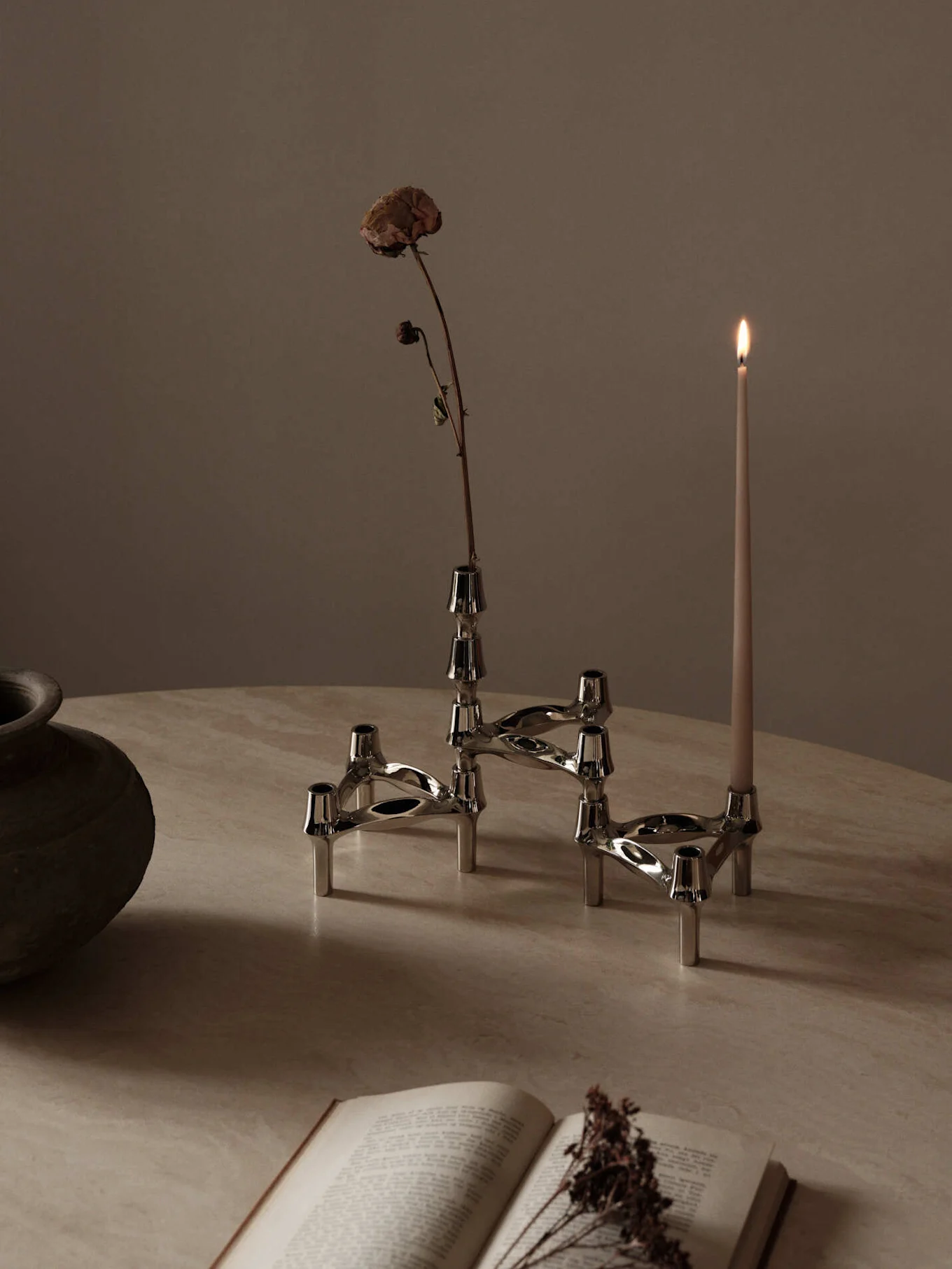 Sistema di candelabri modulare argentato con un fiore essiccato e una candela accesa, su un tavolo chiaro con un libro aperto.