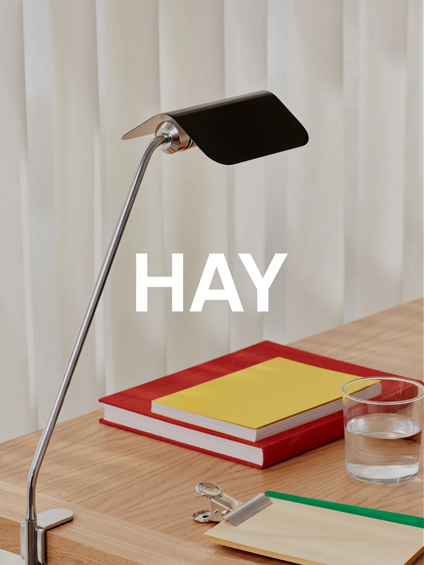 Una lampada da scrivania cromata con paralume nero illumina una scrivania in legno con due libri, un bicchiere d'acqua e un fermacarte.