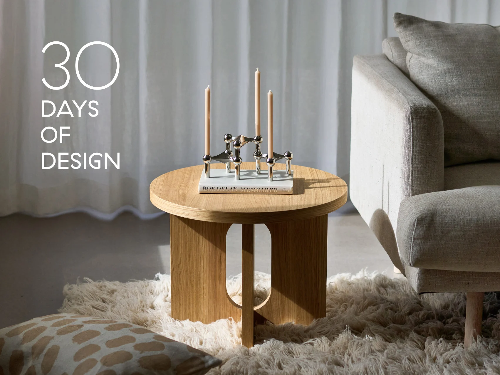 Soggiorno moderno con tavolino rotondo in legno, candelabro d'argento con tre candele, un libro e un divano beige. Testo: '30 GIORNI DI DESIGN'.