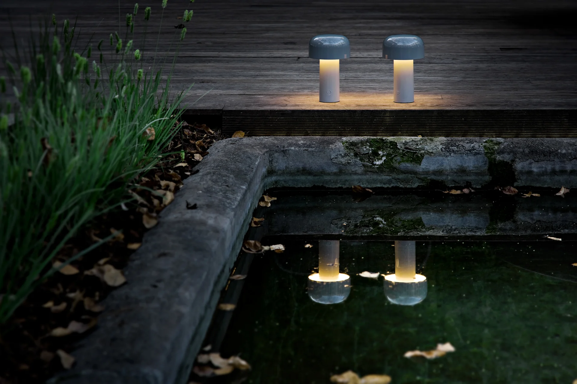 Due lampade da tavolo portatili a forma di fungo con luce gialla calda su un ponte di legno scuro vicino a uno stagno, che riflettono la loro luce.