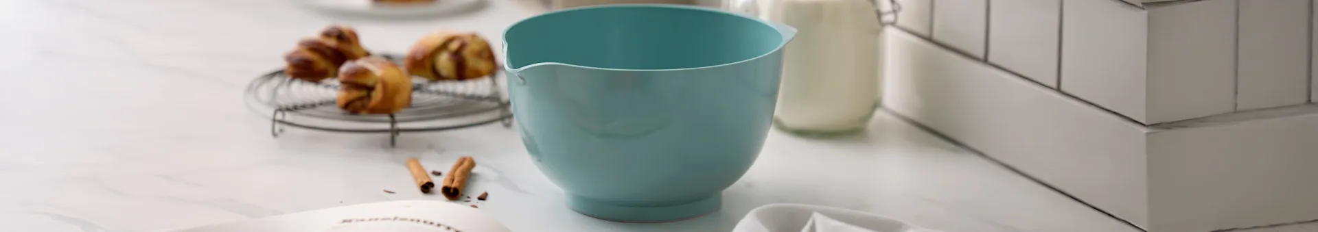 Ciotola azzurra, dolci su griglia, bottiglia di latte e libro di ricette su piano di lavoro in marmo.