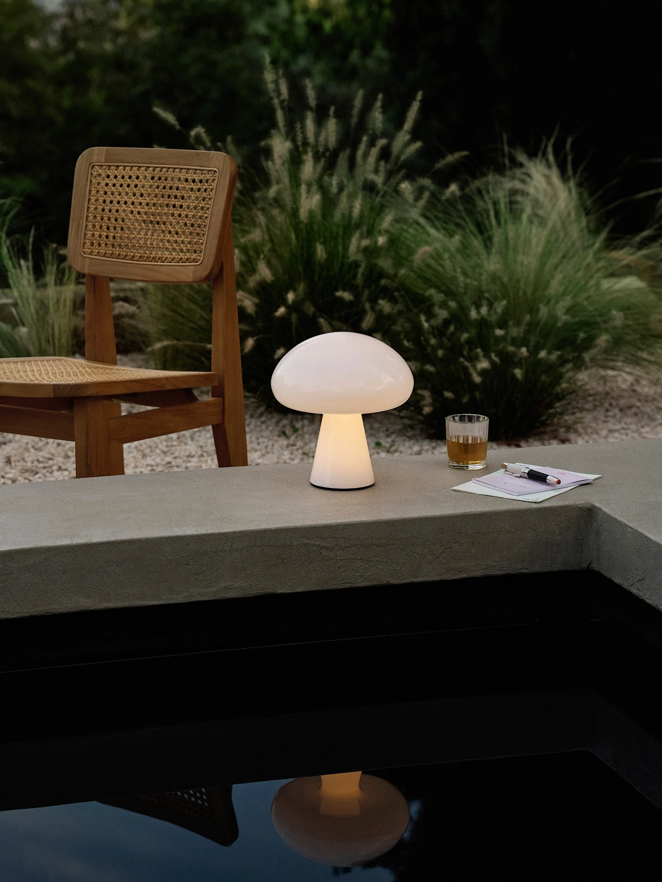Una lampada a forma di fungo luminoso su un bordo di cemento accanto a una piscina, con una sedia di legno, una bevanda e una penna.