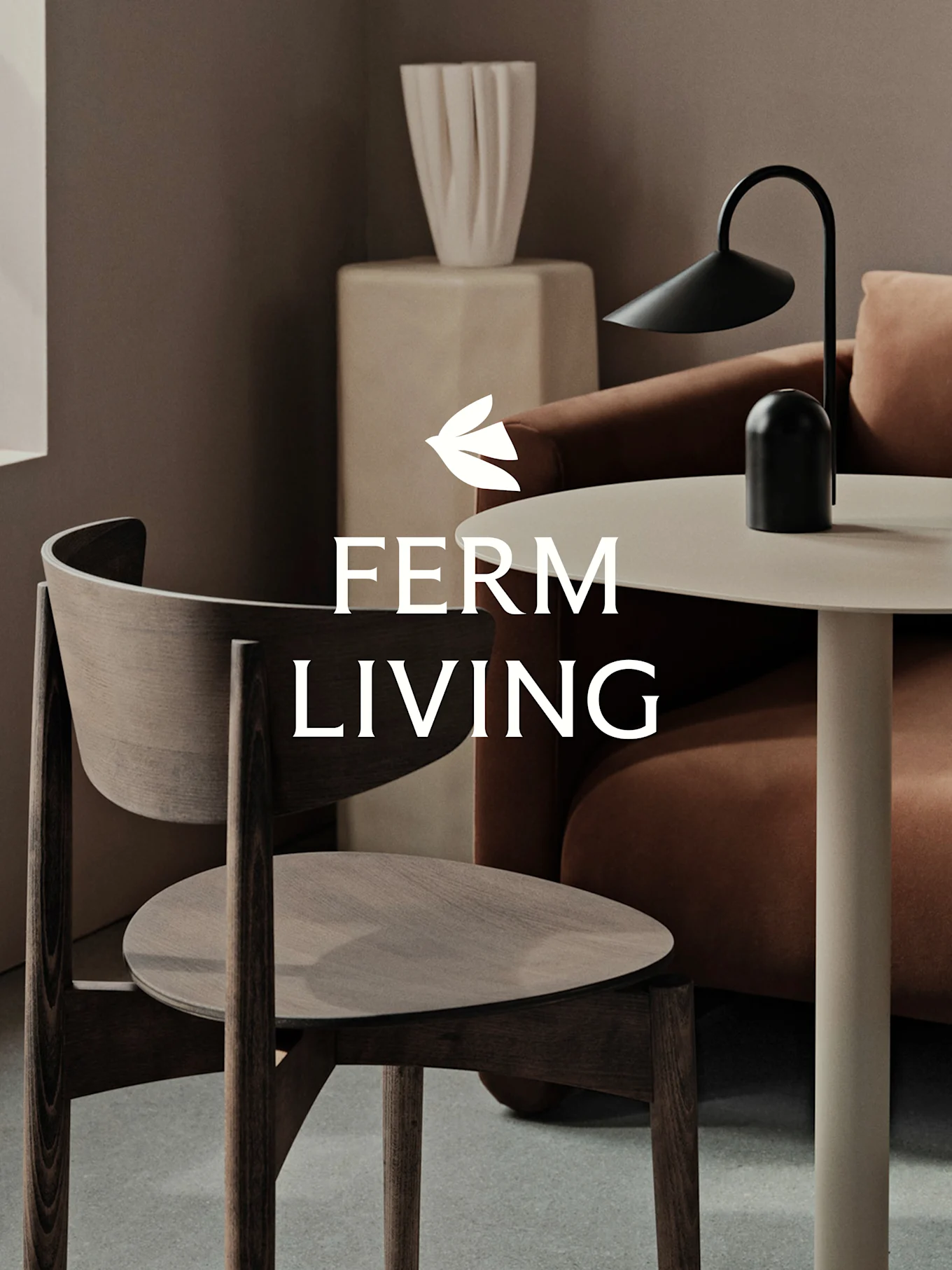 Un moderno spazio abitativo con sedia in legno scuro, tavolino chiaro con lampada nera, divano color terracotta e vaso scultoreo. Logo 'FERM LIVING'.