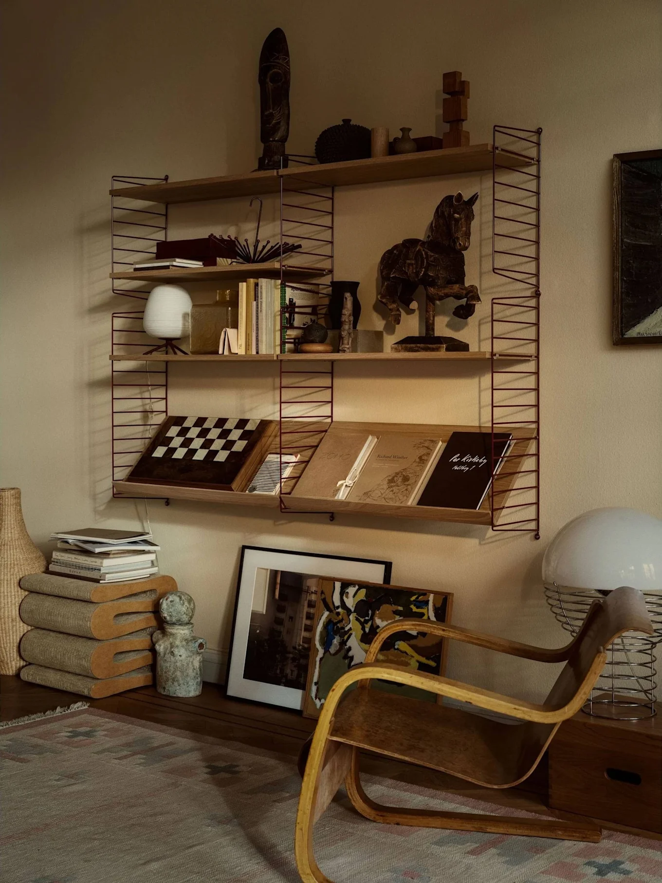 Una stanza accogliente con una libreria piena di libri, arte e decorazioni, accanto a una poltrona in legno curvato e una grande lampada bianca.