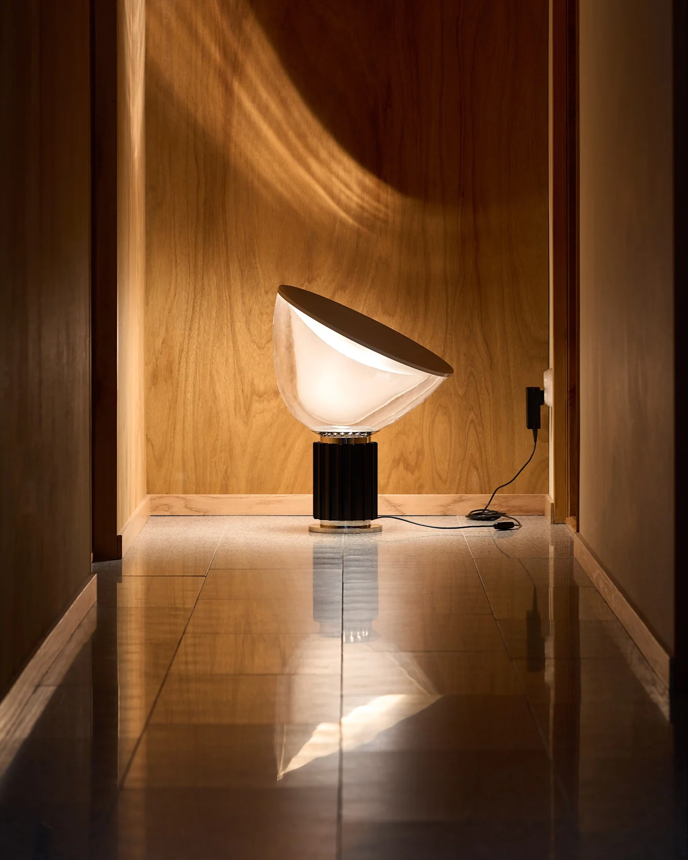 Lampada moderna con paralume bianco opaco semicircolare e base scanalata nera in corridoio rivestito in legno con pavimento riflettente. Luce sulla parete.