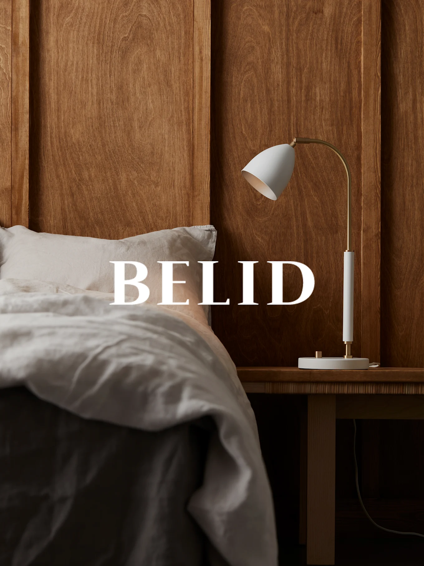 Lampada bianca e ottone su comodino in legno, con la scritta 'BELID' sopra un letto con lenzuola di lino.