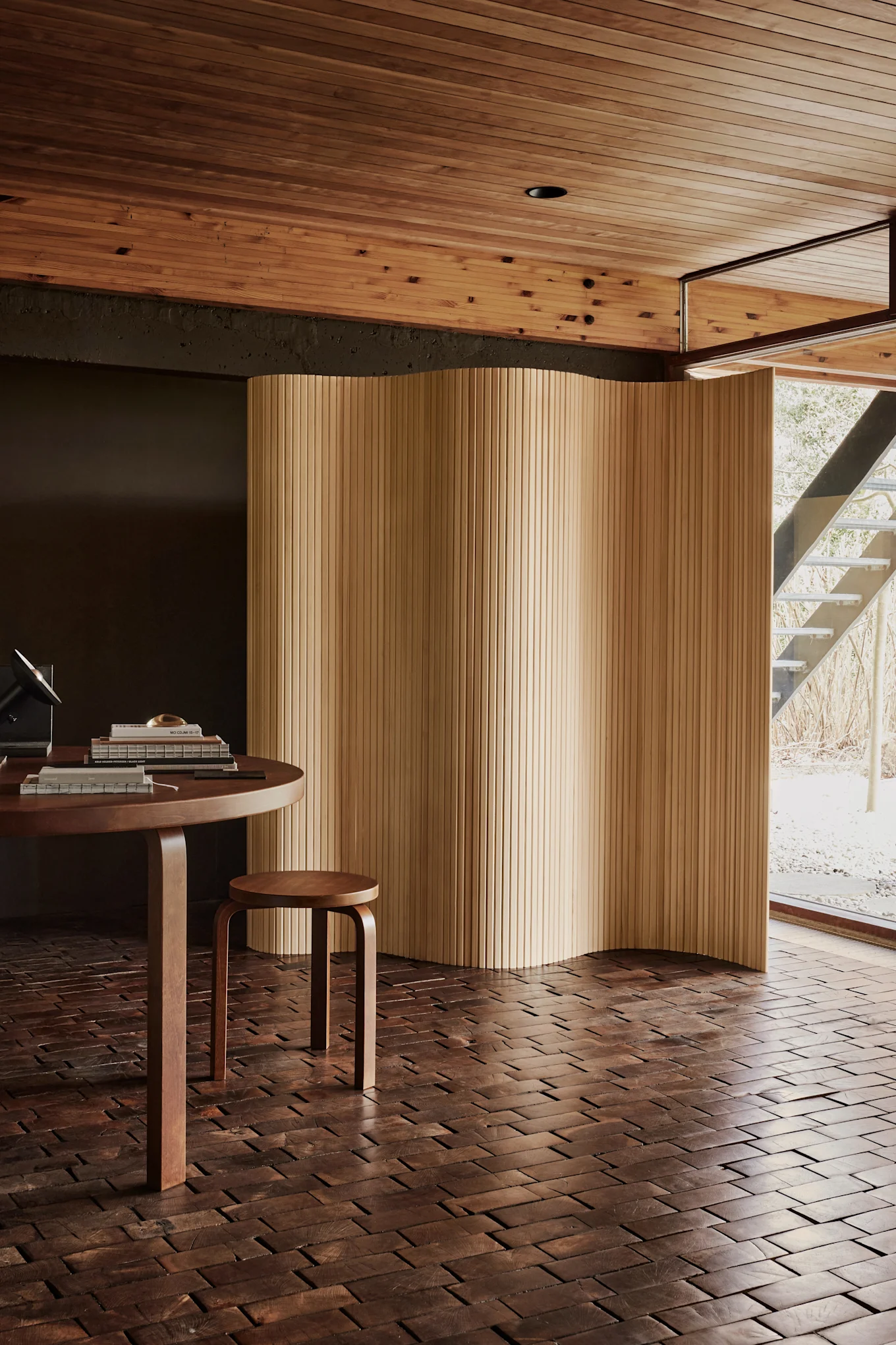 Stanza moderna con tavolo rotondo in legno, sgabello e schermo ondulato in legno. Pavimento a blocchi di legno scuro, soffitto a doghe di legno chiaro e finestra su una scala esterna.