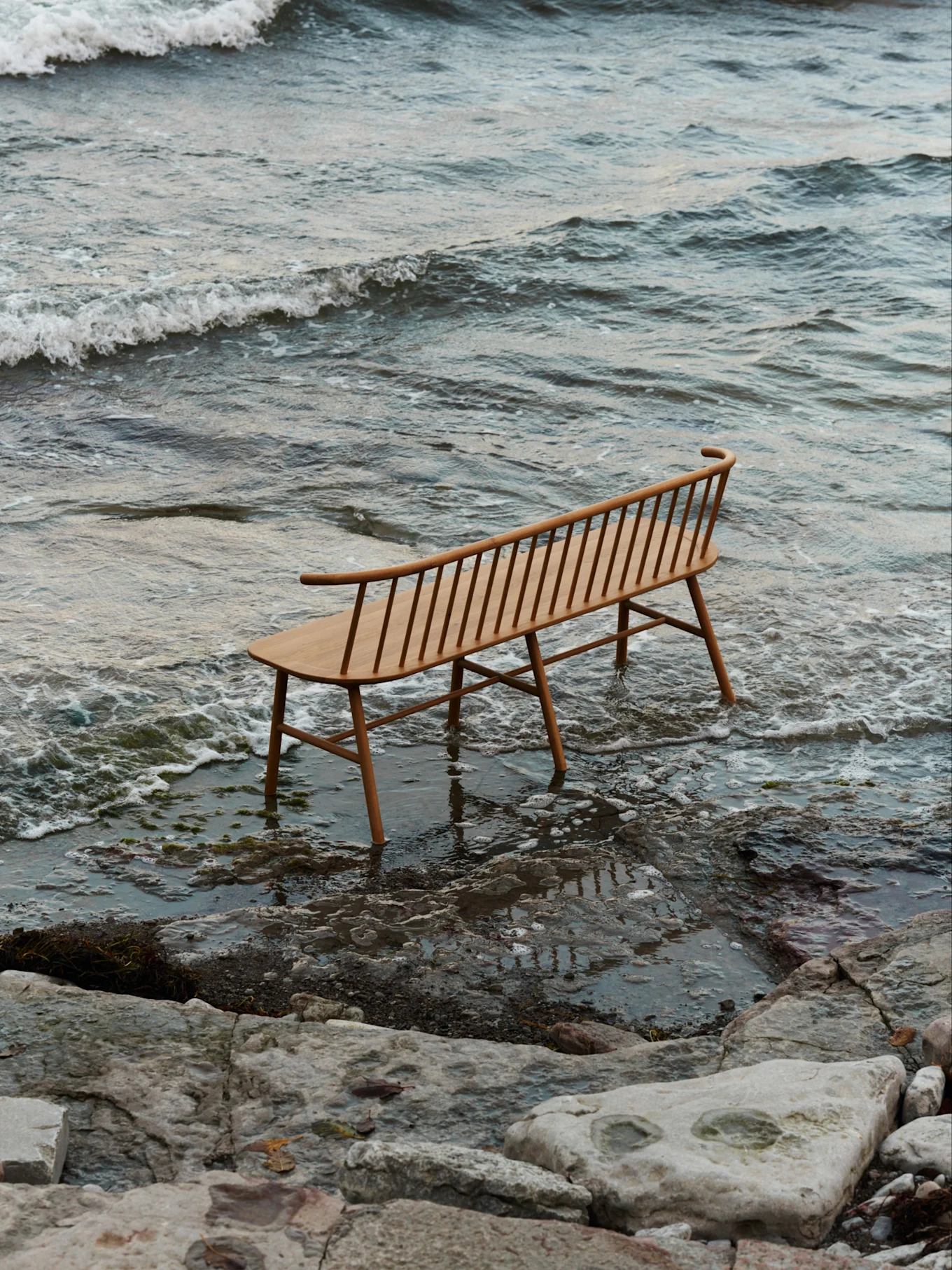 Una panca a doghe di legno chiaro con schienale curvo si trova in acque poco profonde su una spiaggia rocciosa con onde leggere.